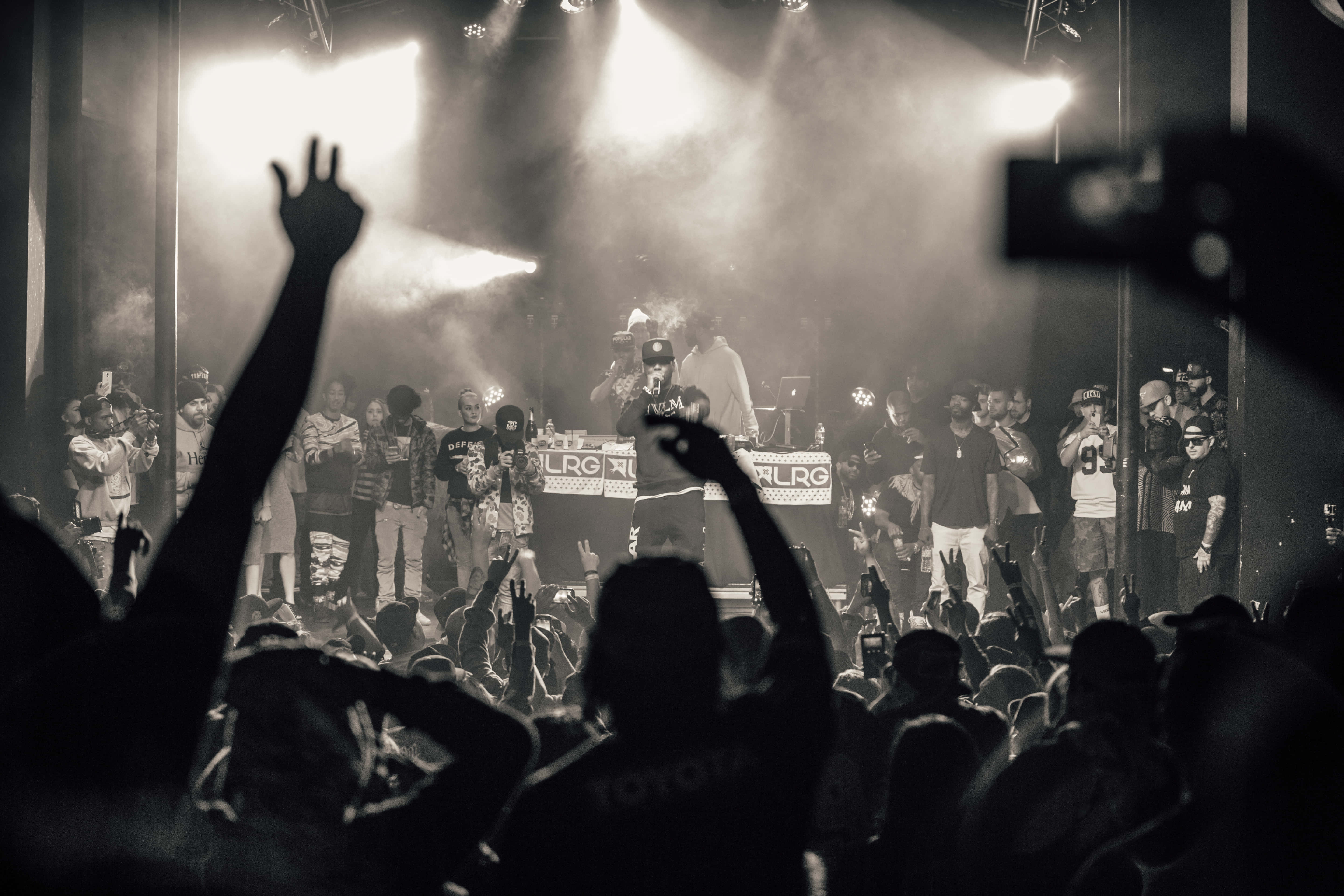 silhouetted concertgoers, smoky black and white venue, lit stage, 5K Desktop Wallpaper