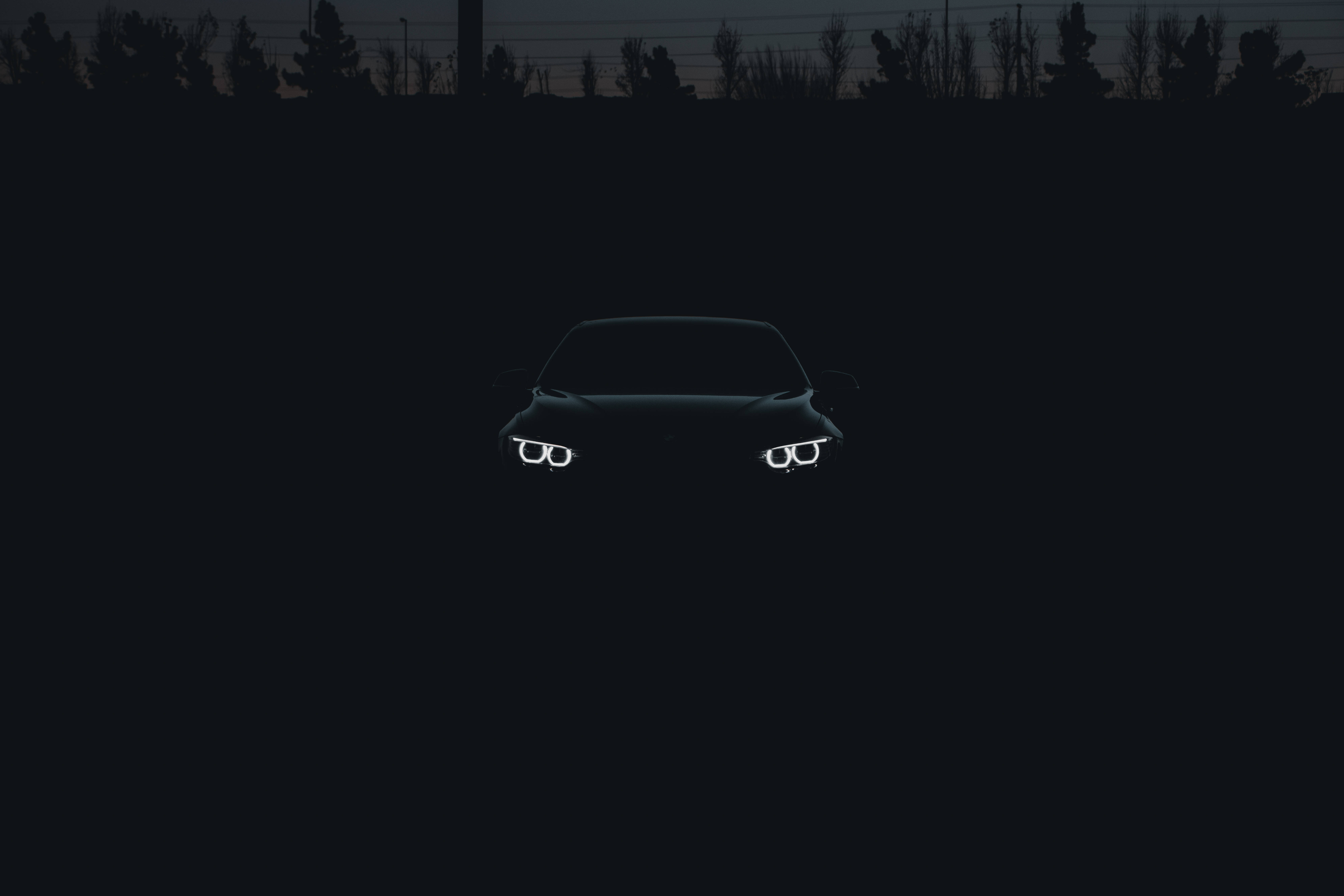 Black BMW, glowing headlights, silhouetted treeline, 5K Desktop Wallpaper