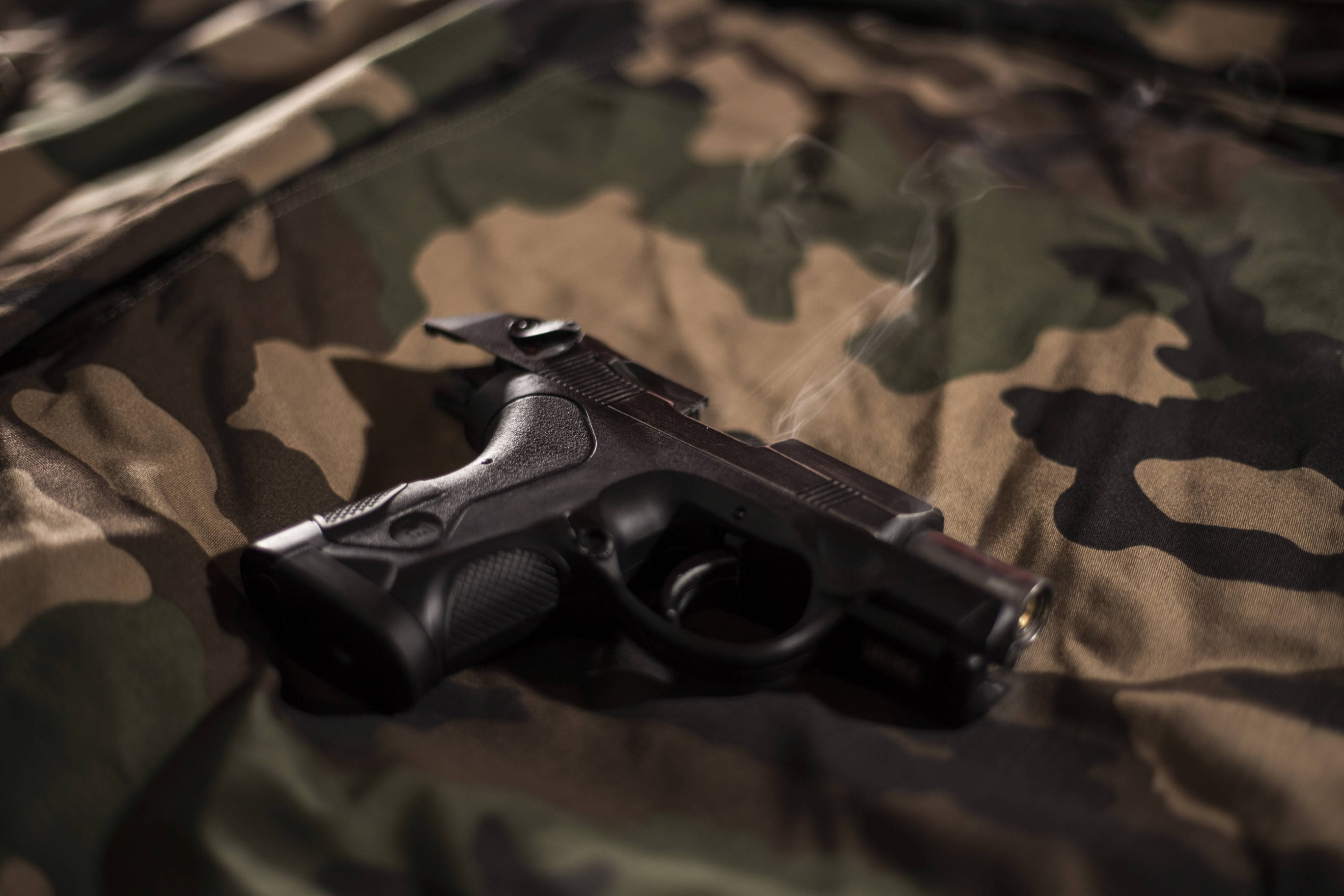 black handgun, camouflage fabric, smoke rising from its barrel, 2K Desktop Wallpaper