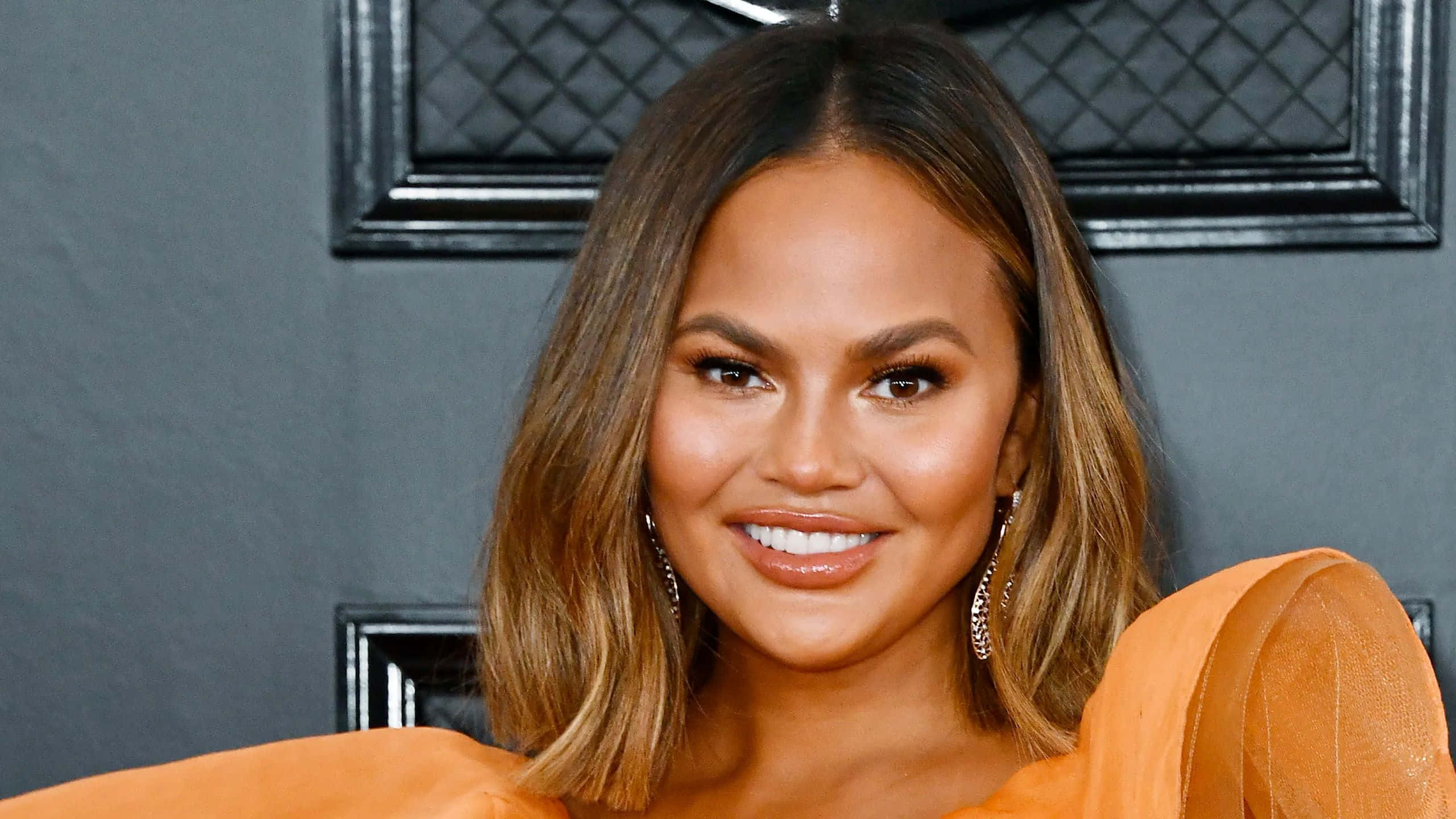 Chrissy Teigen, orange gown, dark gray textured wall, 2K Desktop Wallpaper