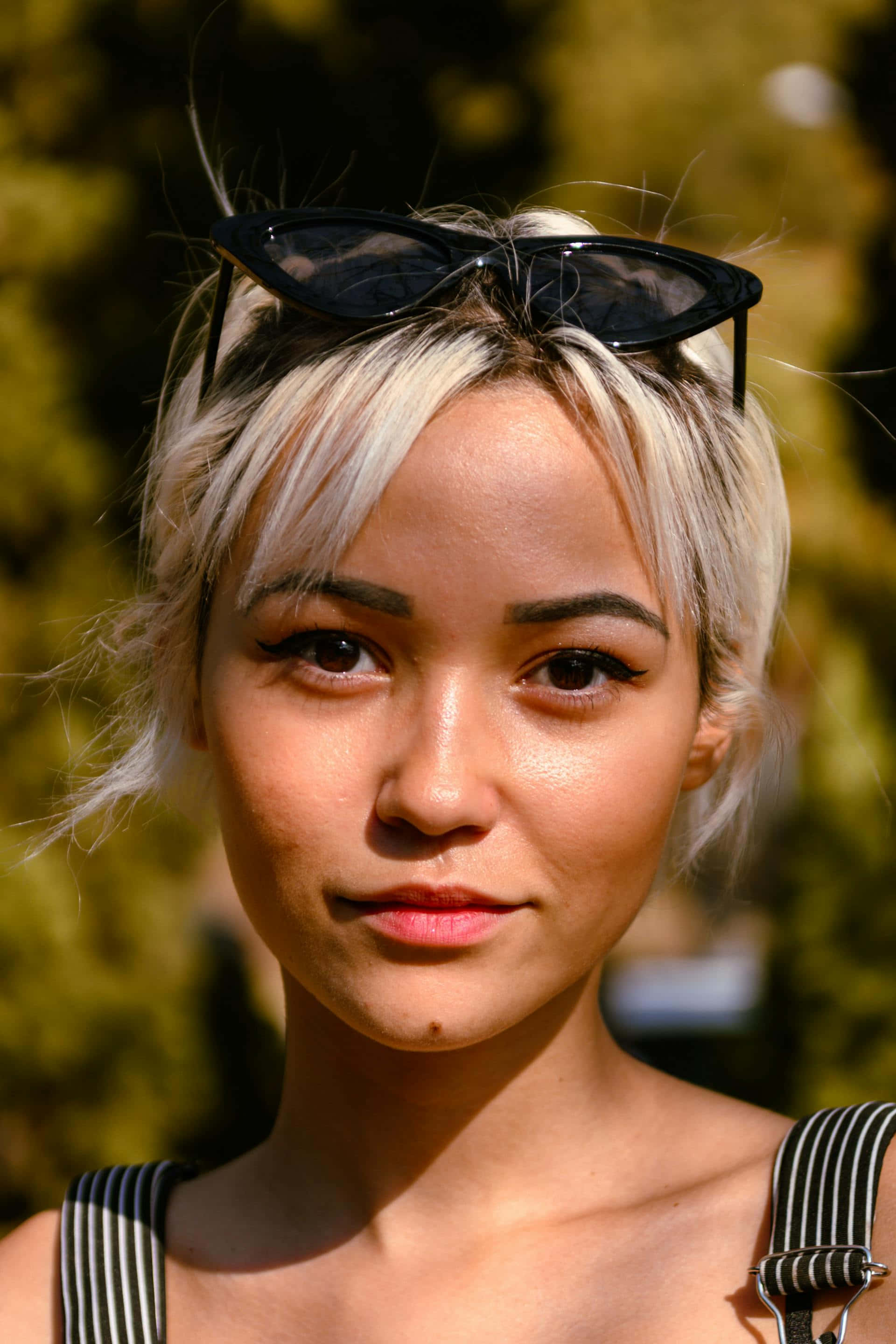 platinum blonde hair, black cat-eye sunglasses, sunlit green foliage background, 2K Phone Wallpaper