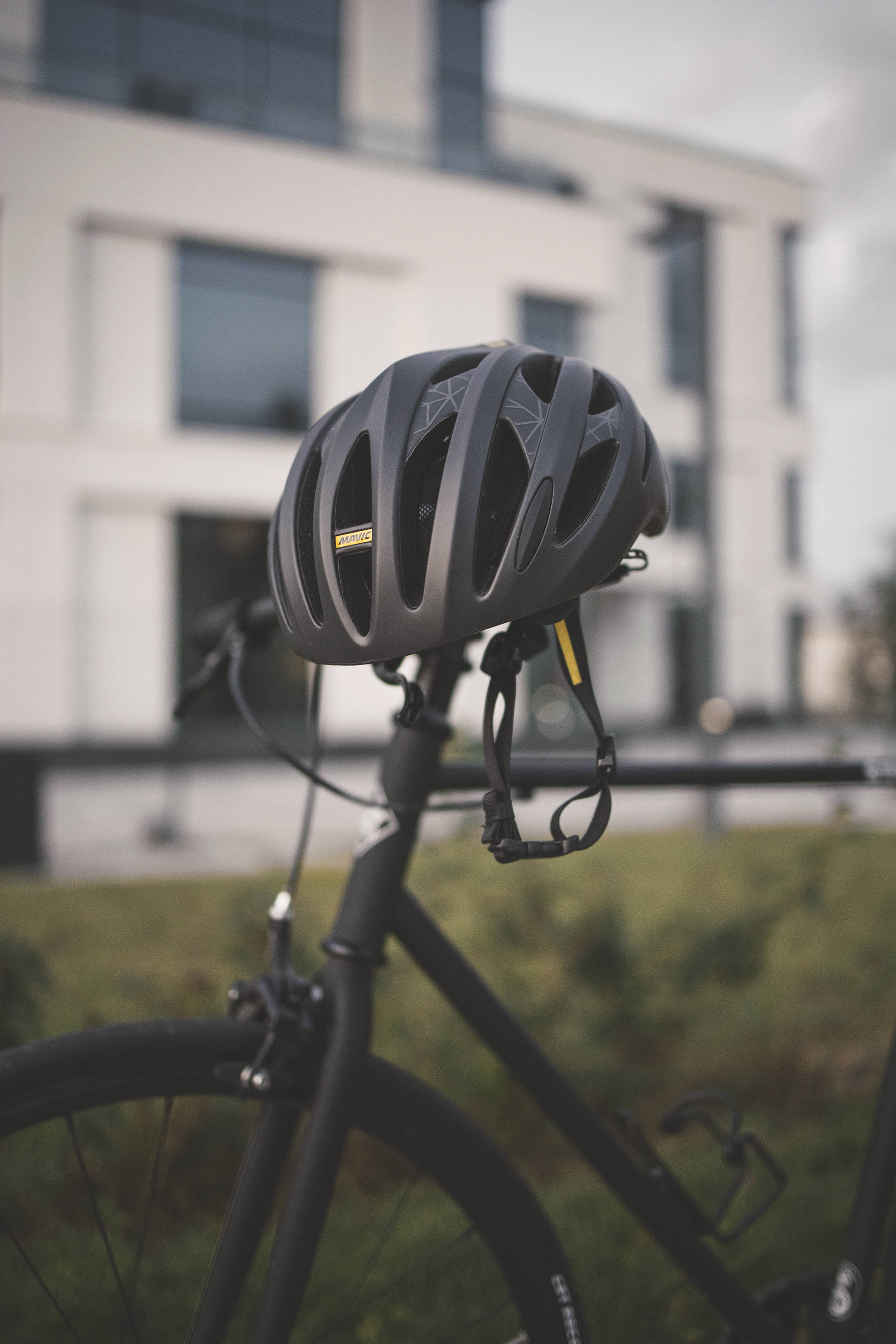 matte black cycling helmet, sleek black bike, blurred modern building backdrop, 2K Phone Wallpaper