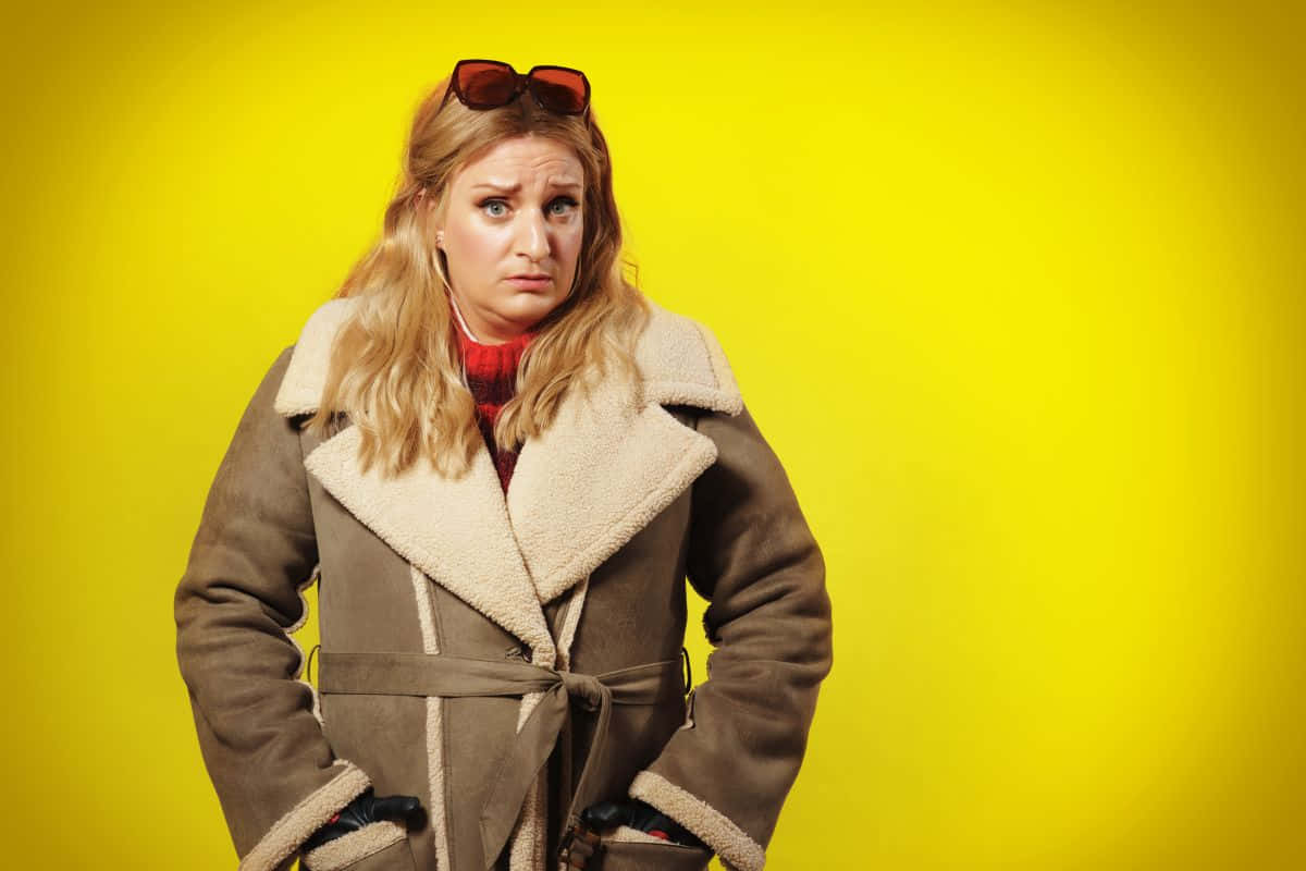 Daisy May Cooper, brown shearling coat, bright yellow background, SD Desktop Wallpaper