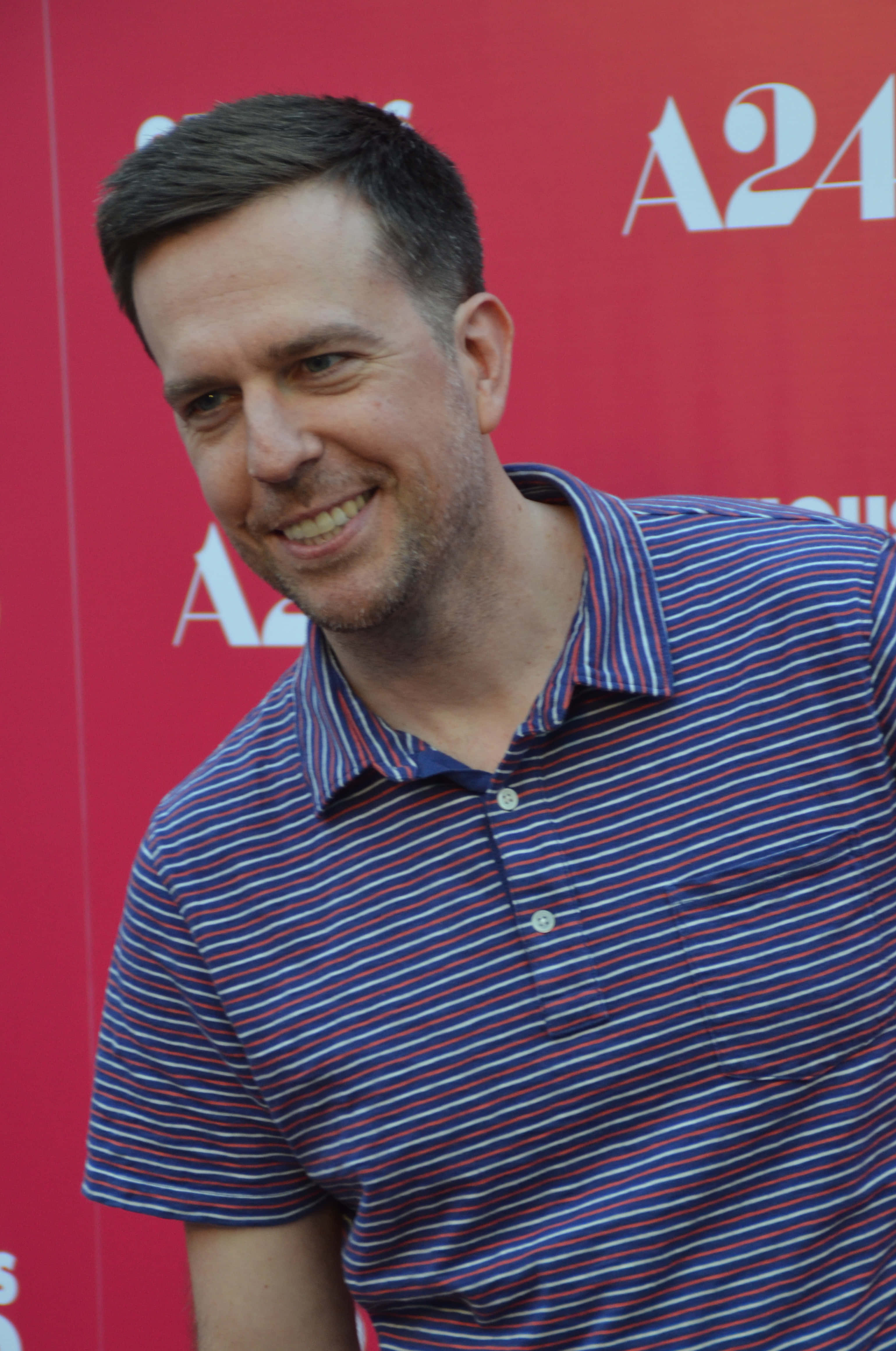 Ed Helms, striped polo shirt, red A24 branded backdrop, 4K Phone Wallpaper