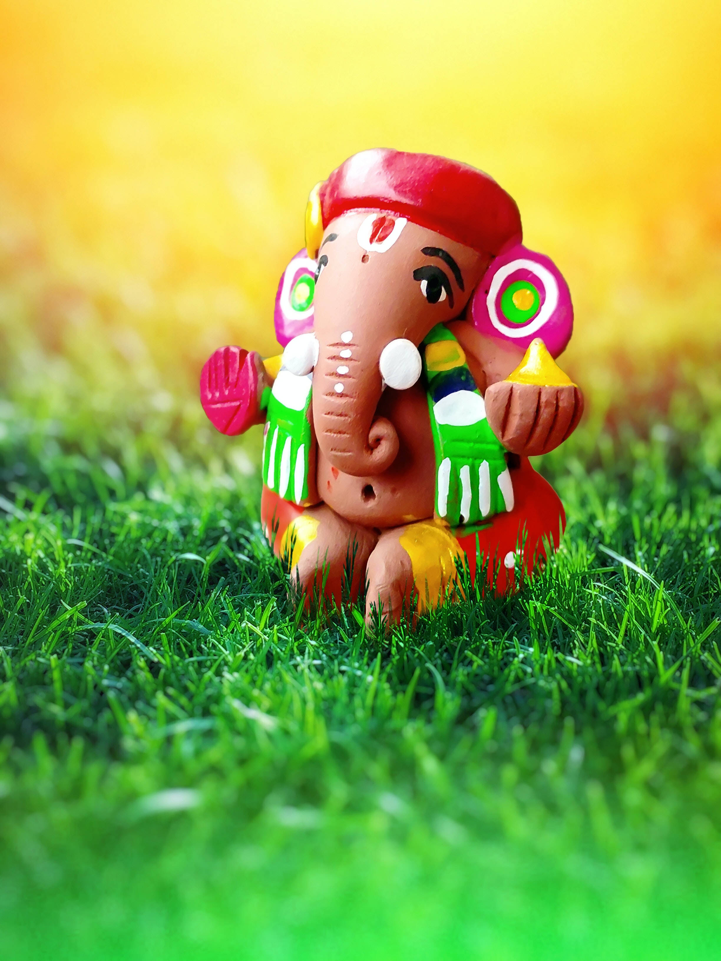clay Ganesha figurine, vibrantly painted, lush green grass, 2K Phone Wallpaper