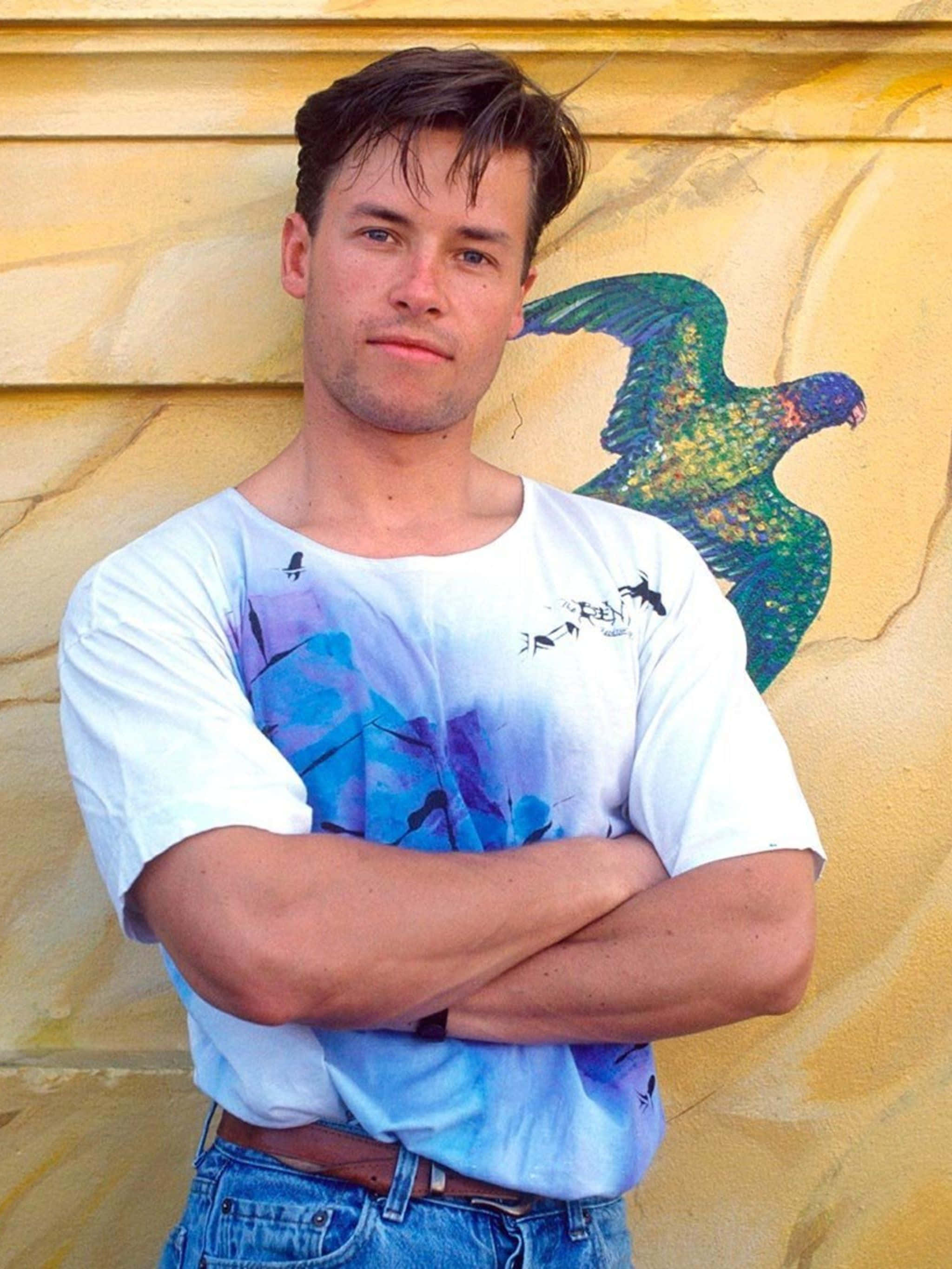 Guy Pearce, textured yellow wall, vibrant green and blue bird mural, 2K Phone Wallpaper