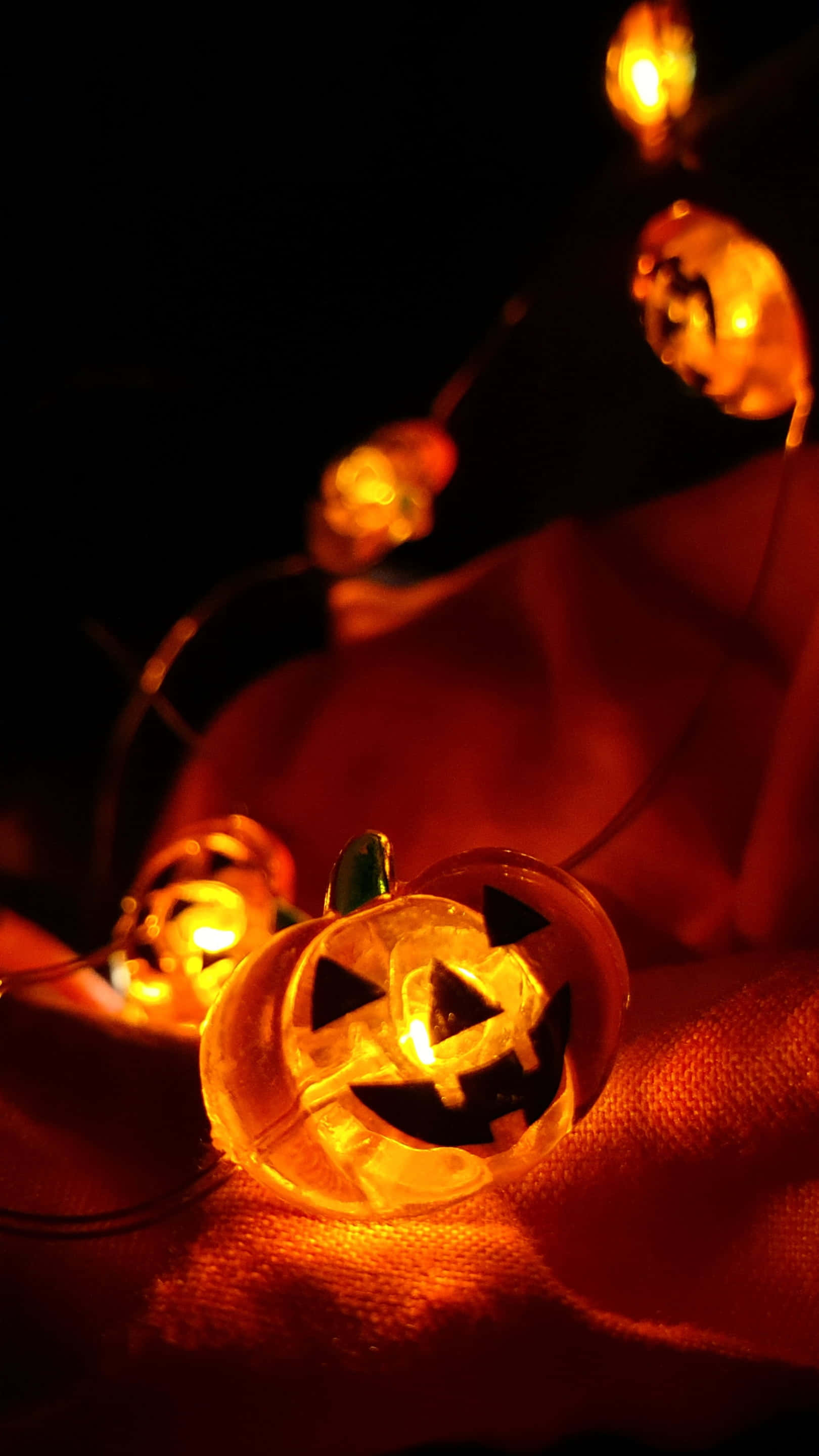jack-o'-lantern string lights, warm orange light, textured red fabric, 2K Phone Wallpaper