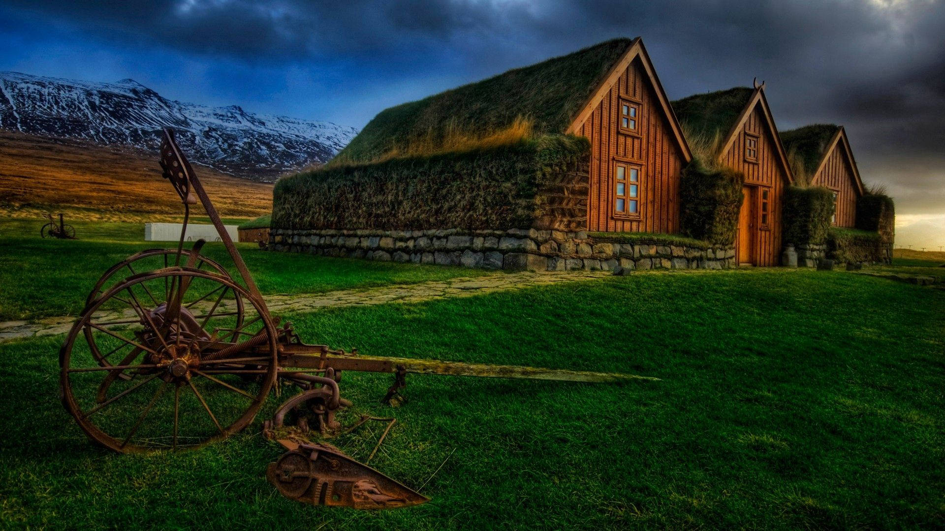 rustic grass-roofed wooden houses, snow-capped mountains, dramatic twilight sky, Full HD Desktop Wallpaper