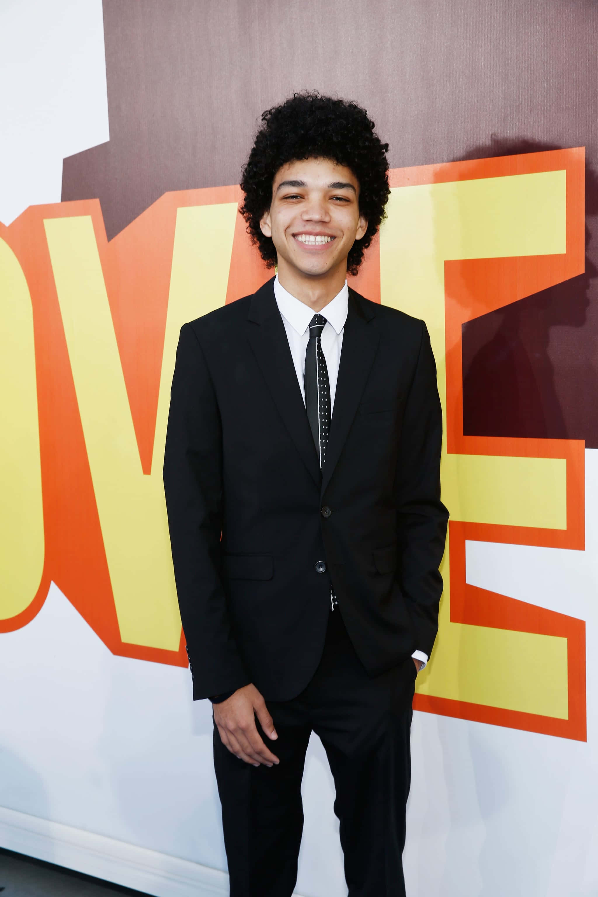 Justice Smith, black suit, bold orange and yellow 'LOVE' backdrop, 2K Phone Wallpaper