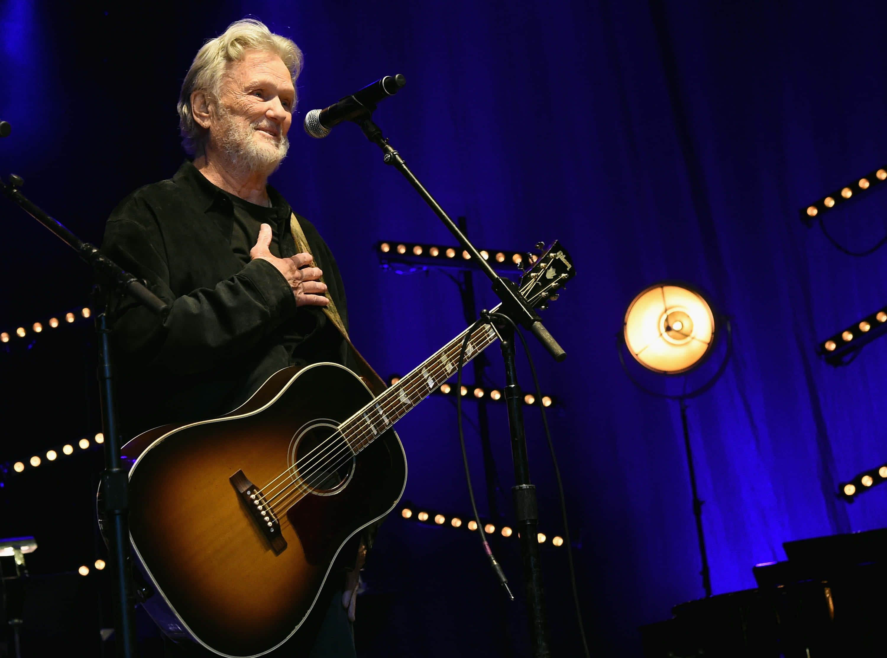 Kris Kristofferson, acoustic guitar, dimly lit stage, 2K Desktop Wallpaper