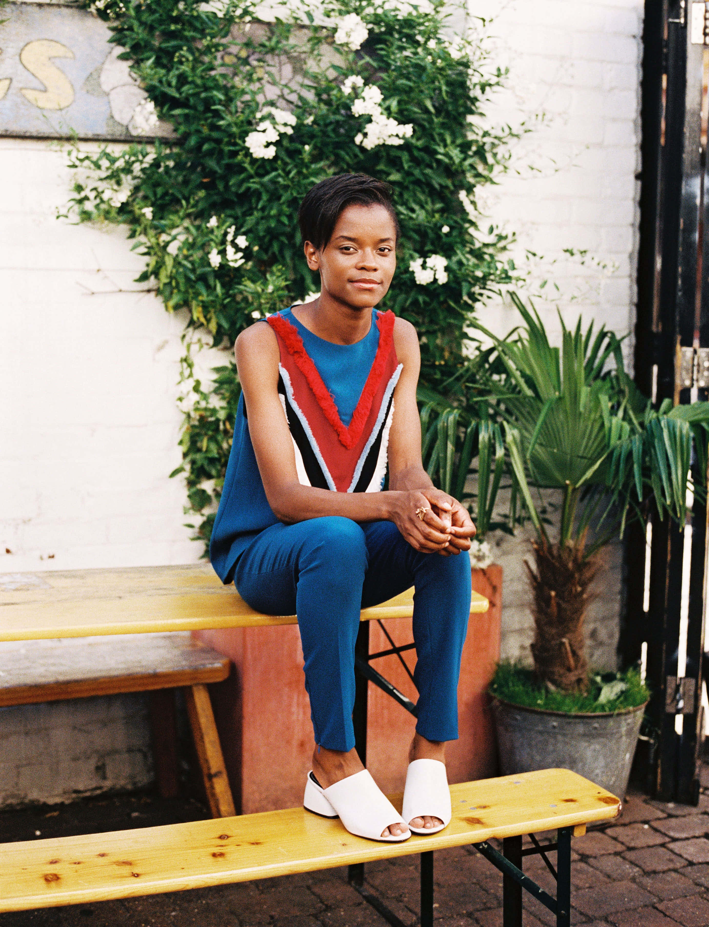 Letitia Wright, bold blue and red geometric top, wooden bench, 2K Phone Wallpaper