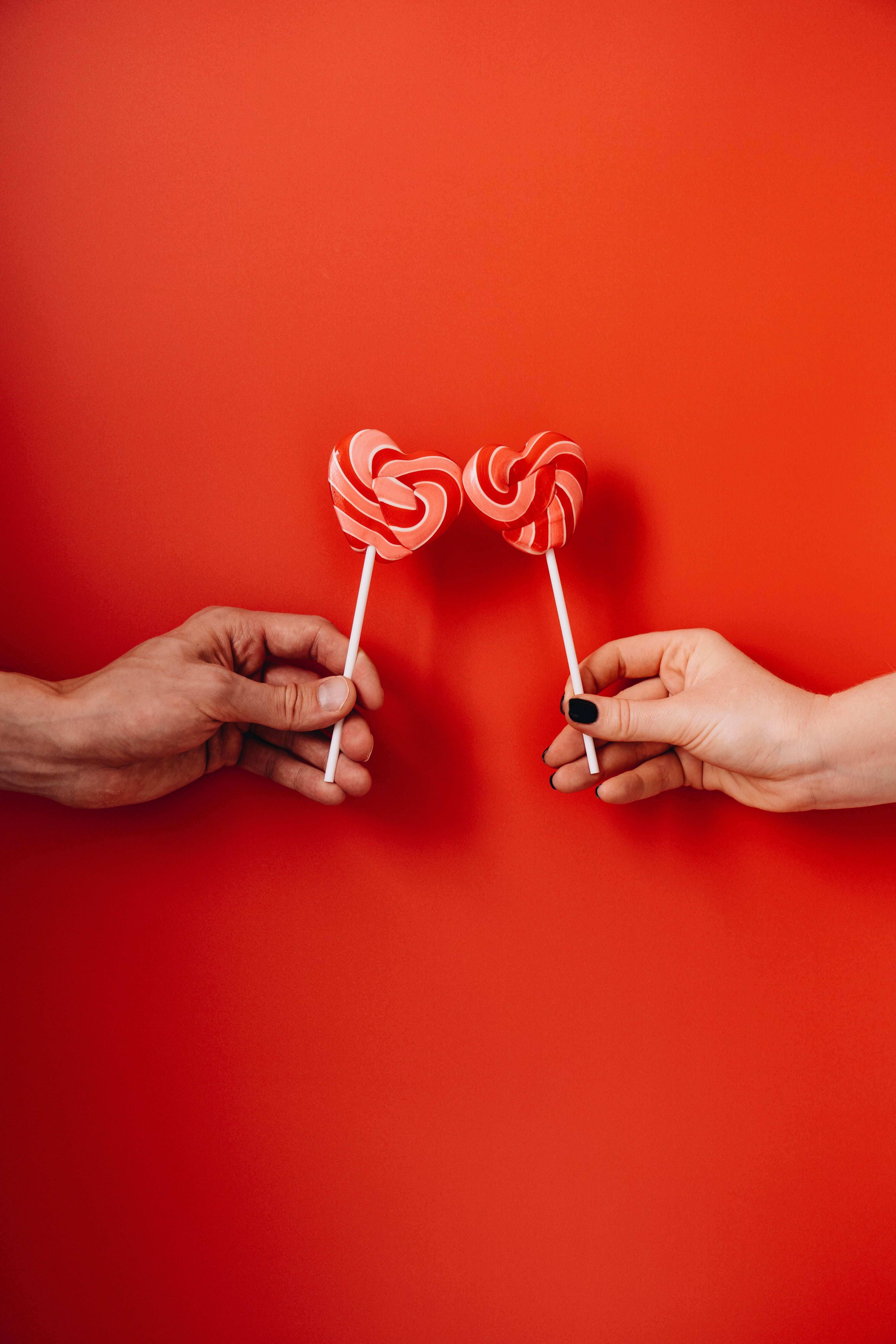 heart-shaped lollipops, vibrant red background, two hands, 5K Phone Wallpaper