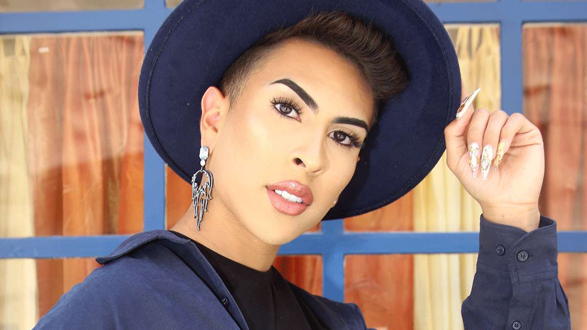 Gay Latino MUA, navy hat, blue-framed wooden backdrop, SD Desktop Wallpaper