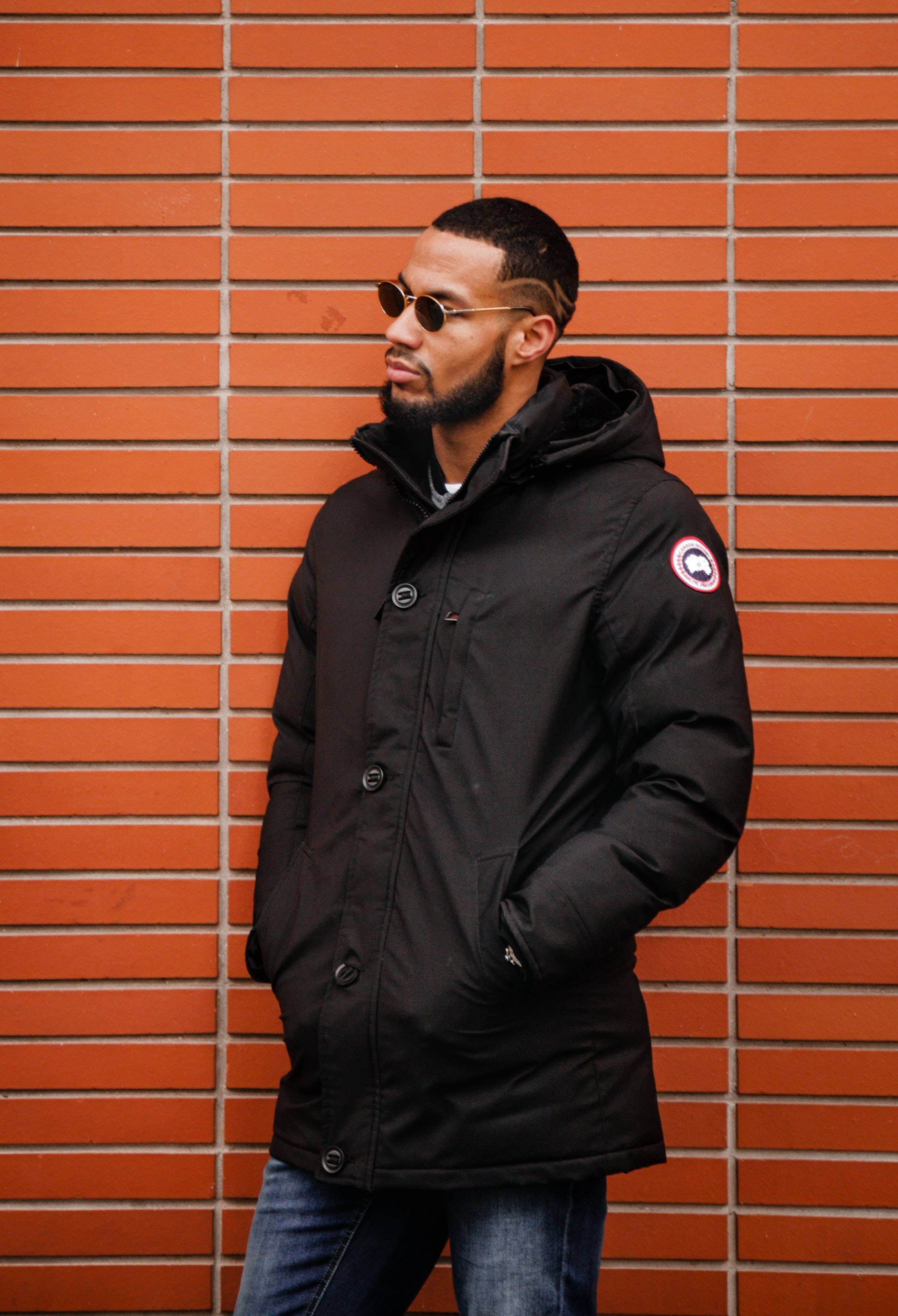 black Canada Goose jacket, warm orange brick wall, sunglasses, 2K Phone Wallpaper