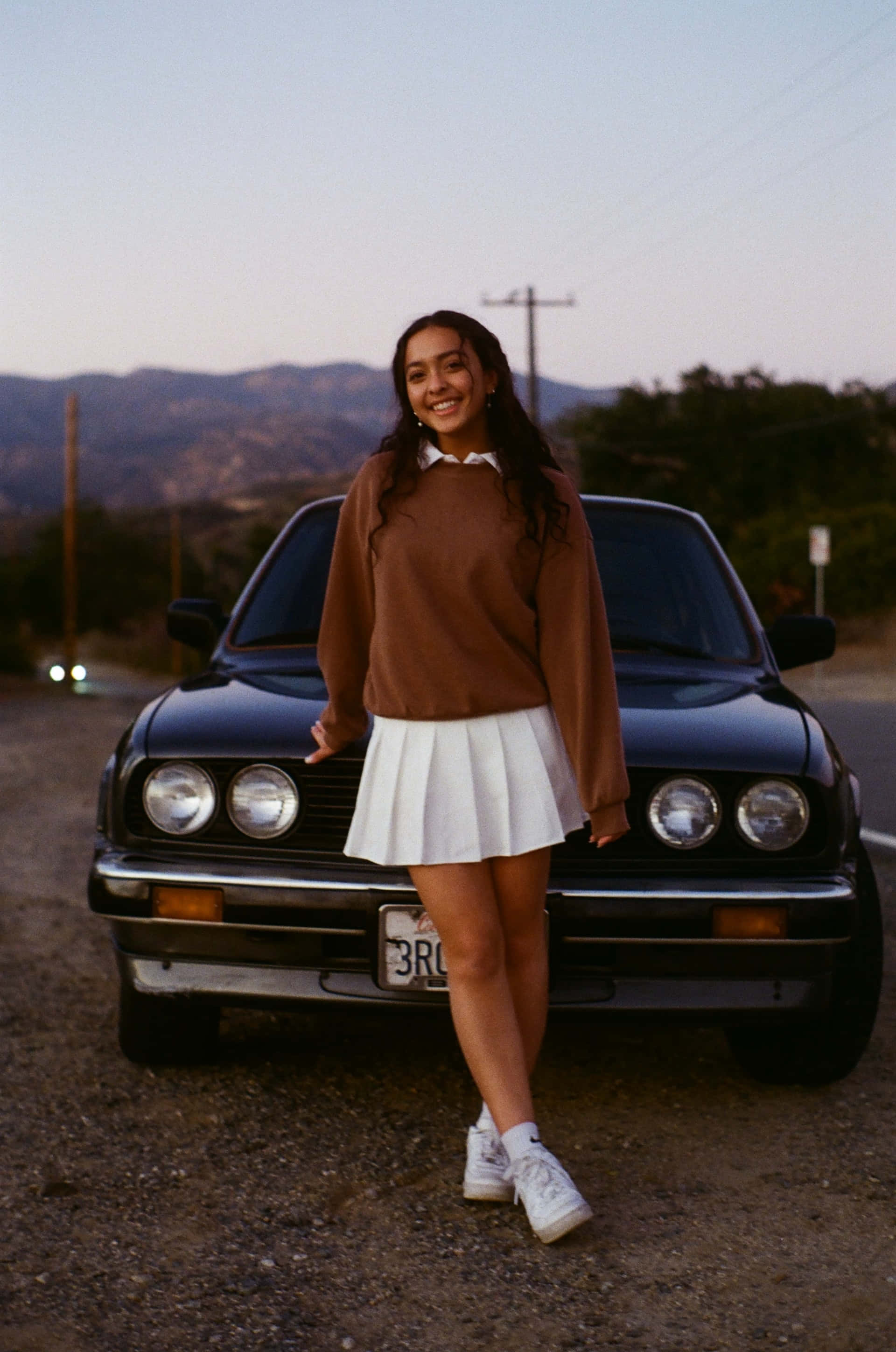 white pleated miniskirt, vintage black car, gravel roadside, 2K Phone Wallpaper
