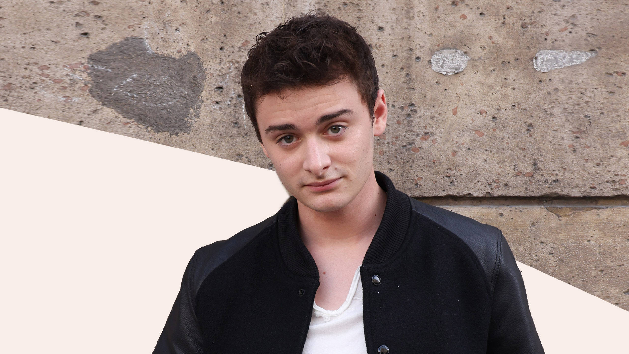 Noah Schnapp, black varsity jacket, textured concrete wall, 2K Desktop Wallpaper