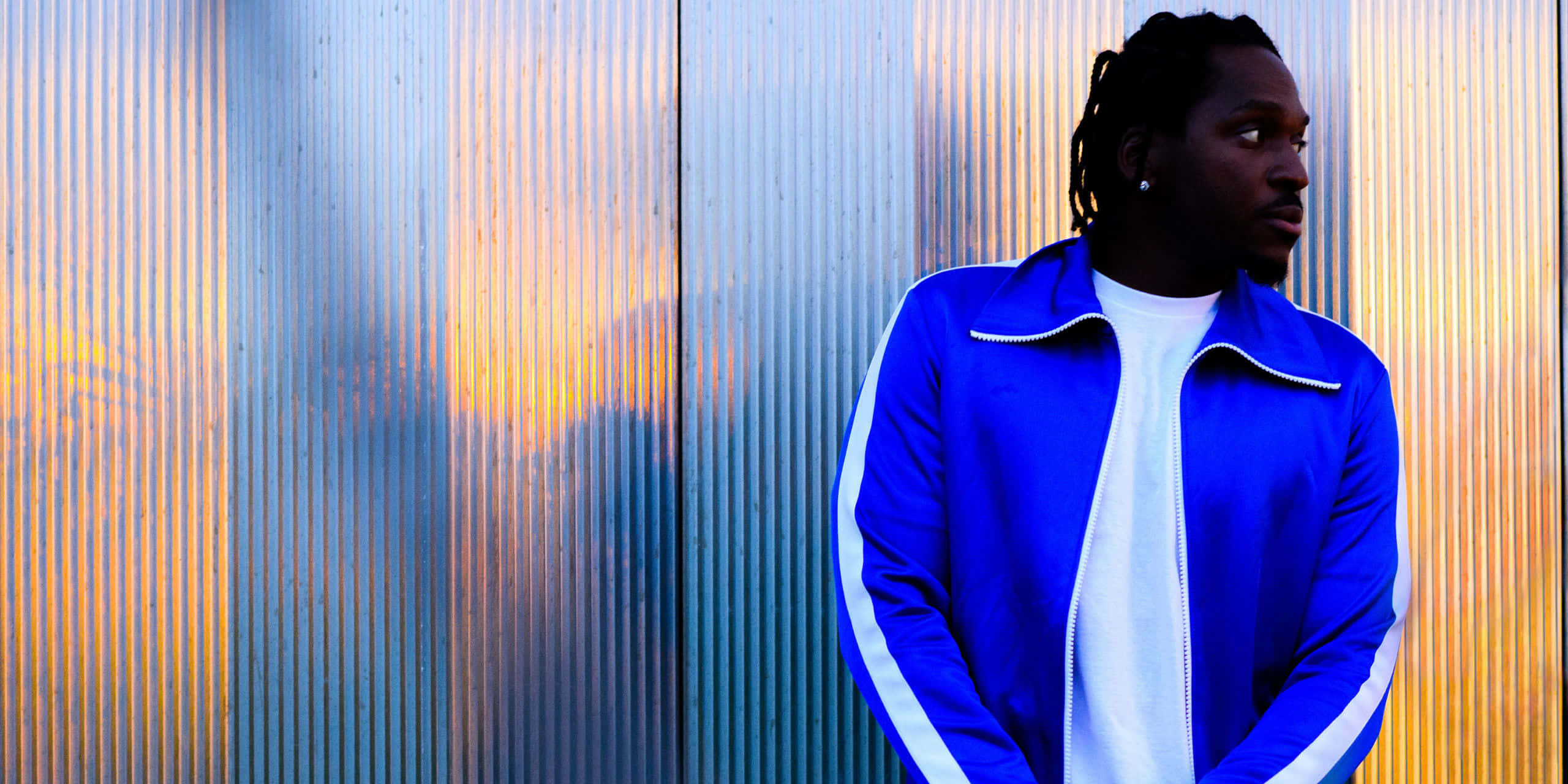 Pusha T, blue track jacket, corrugated metal wall, 2K Desktop Wallpaper