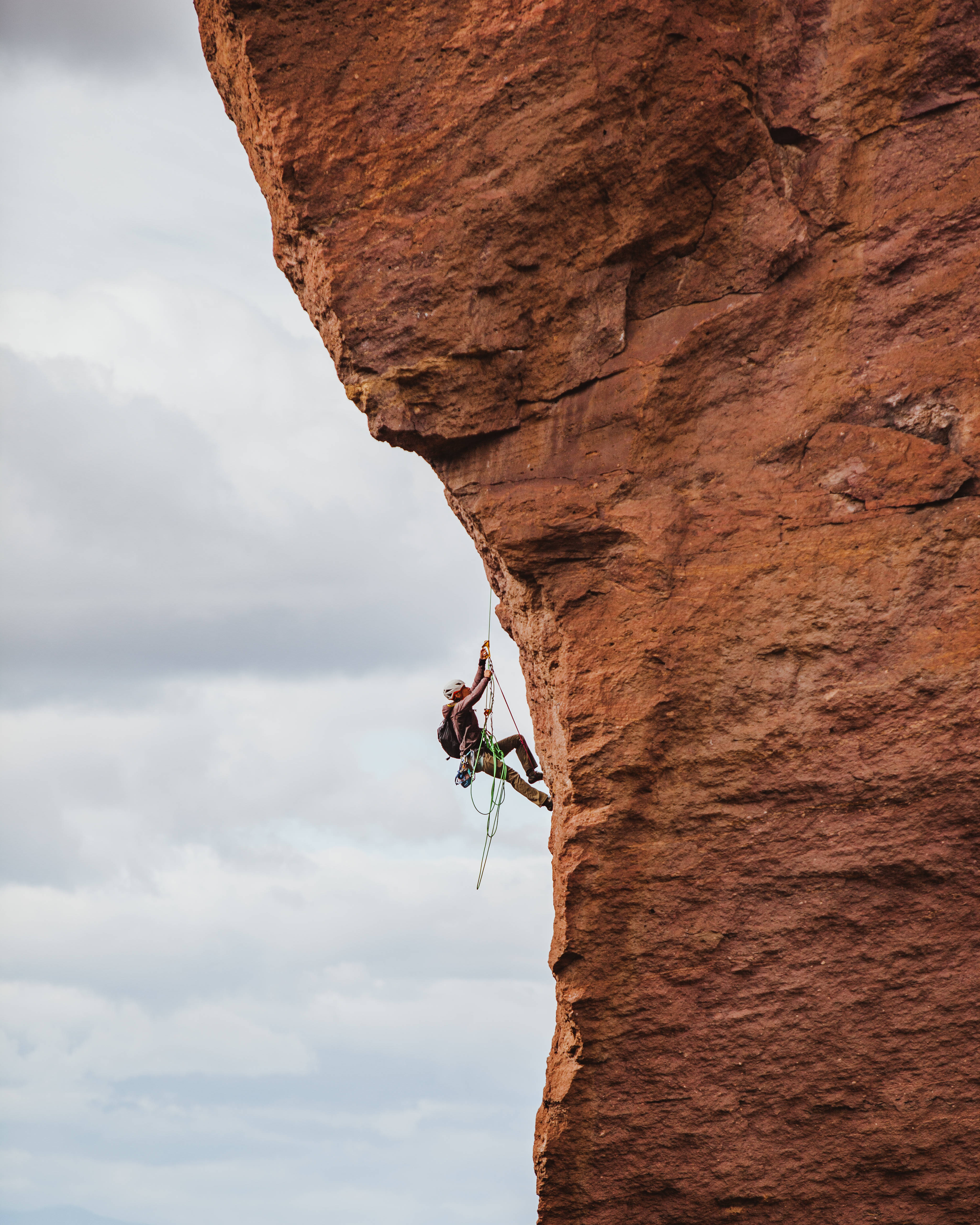 man rock climbing, steep red rock cliff face, cloudy sky, 4K Phone Wallpaper
