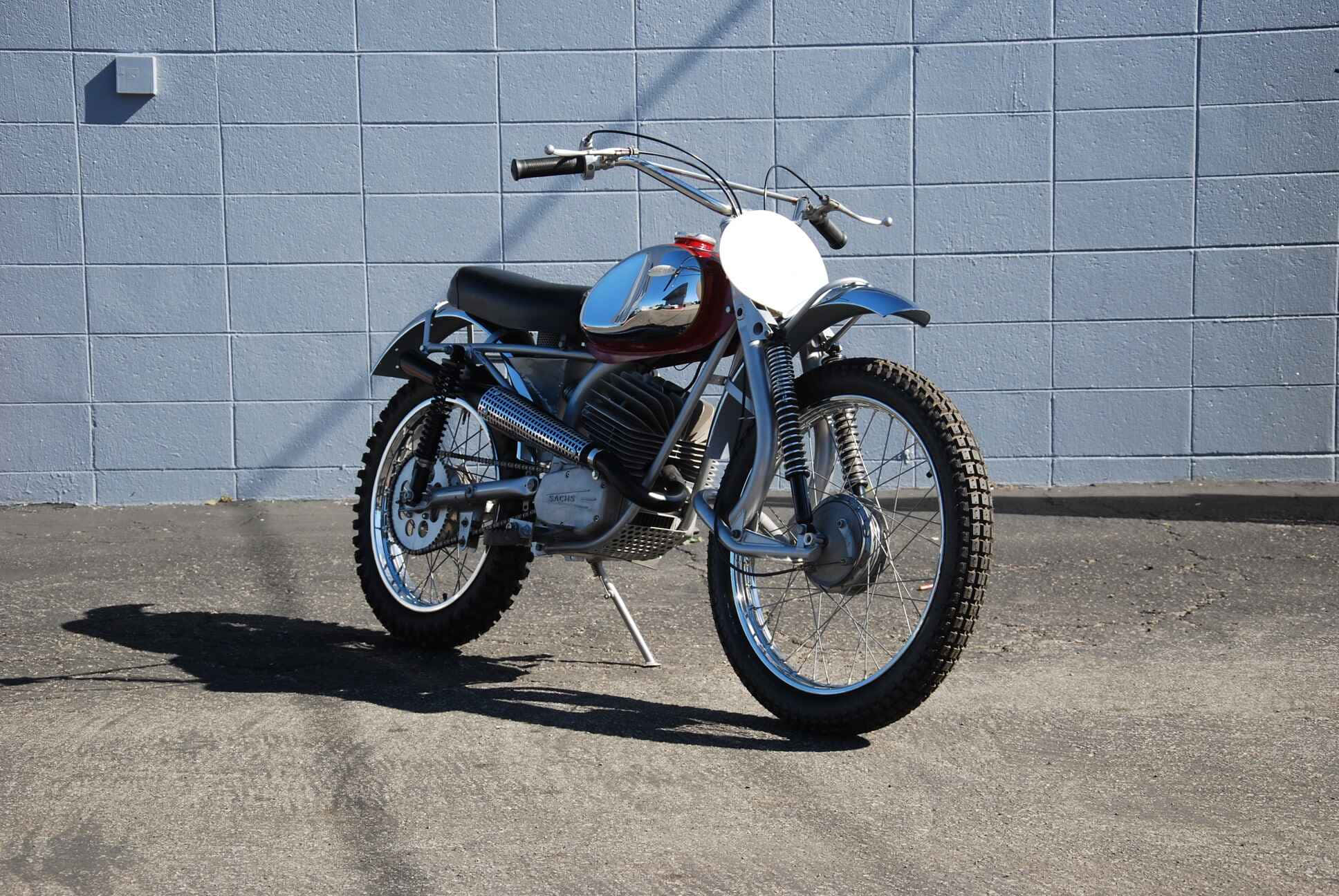 A vintage chrome and red dirt bike stands poised on a concrete lot against a gray cinder block wall under bright sunlight., Full HD Desktop Wallpaper