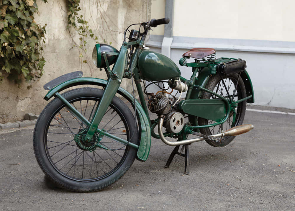 A vintage green motorcycle with chrome accents stands parked on asphalt beside a ivy-covered wall., SD Desktop Wallpaper