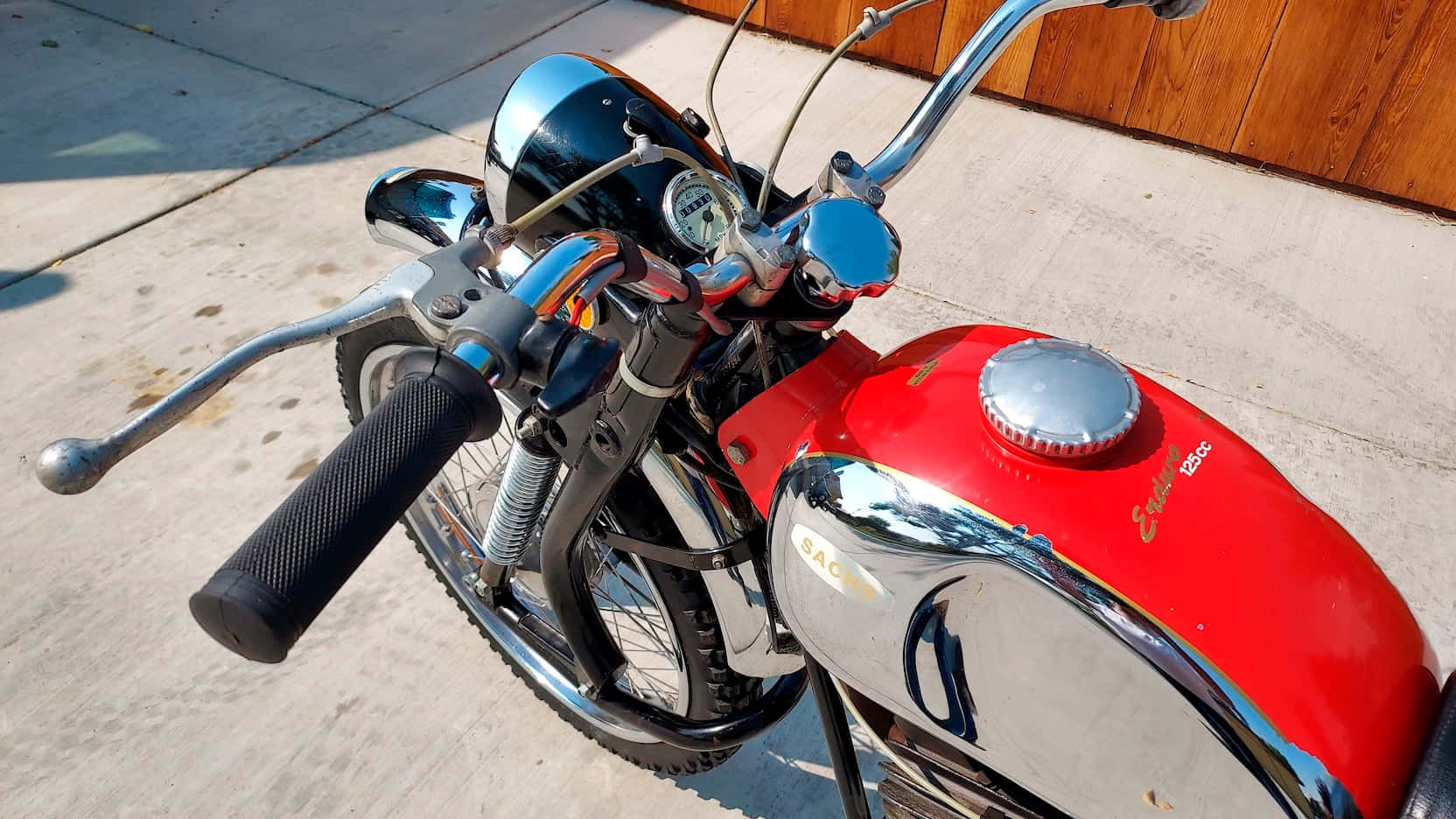 A vintage red and chrome motorcycle with black grips parked on a concrete driveway under bright sunlight., HD Desktop Wallpaper