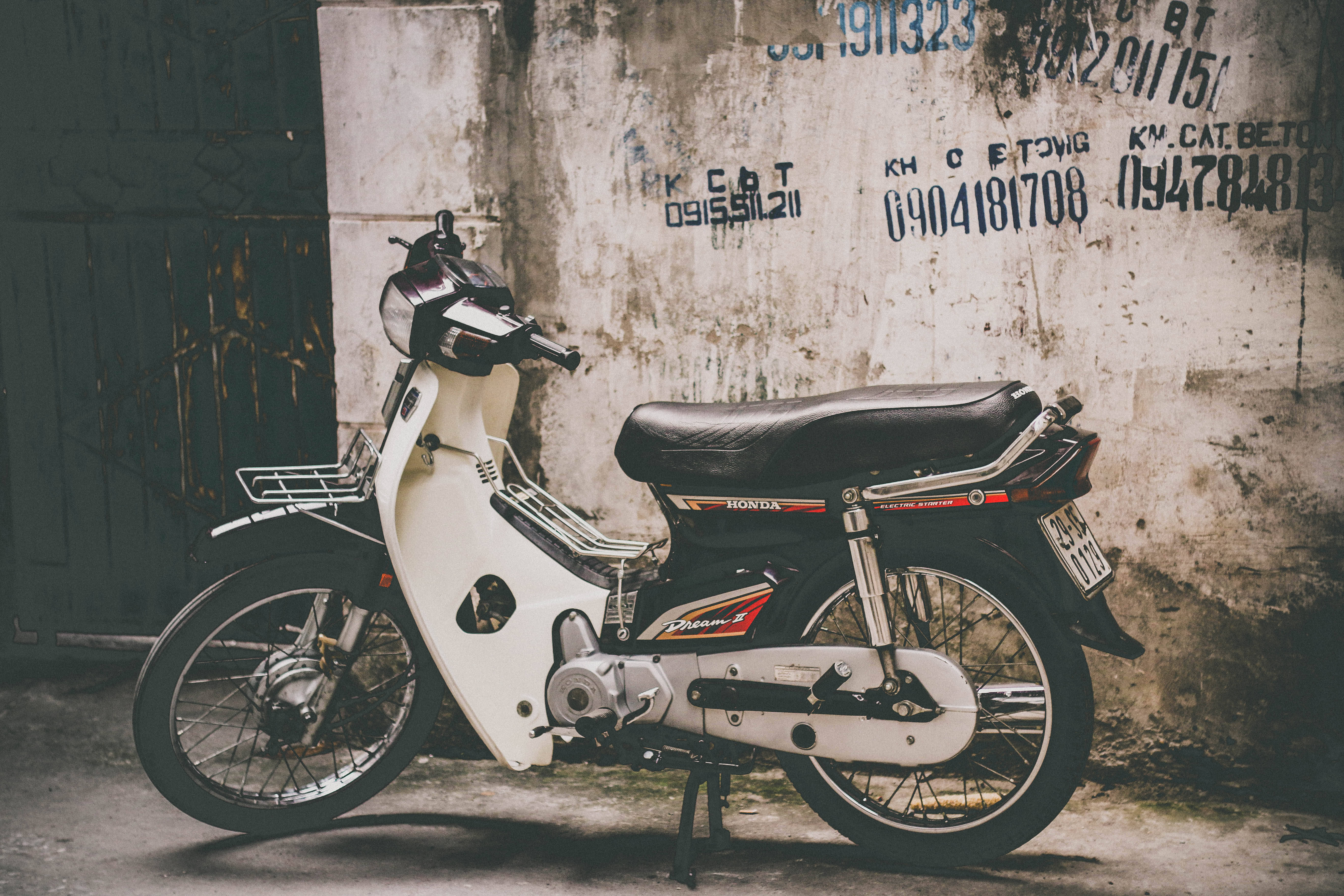 A vintage white Honda Dream motorcycle leans against a weathered concrete wall covered in faded graffiti., 5K Desktop Wallpaper