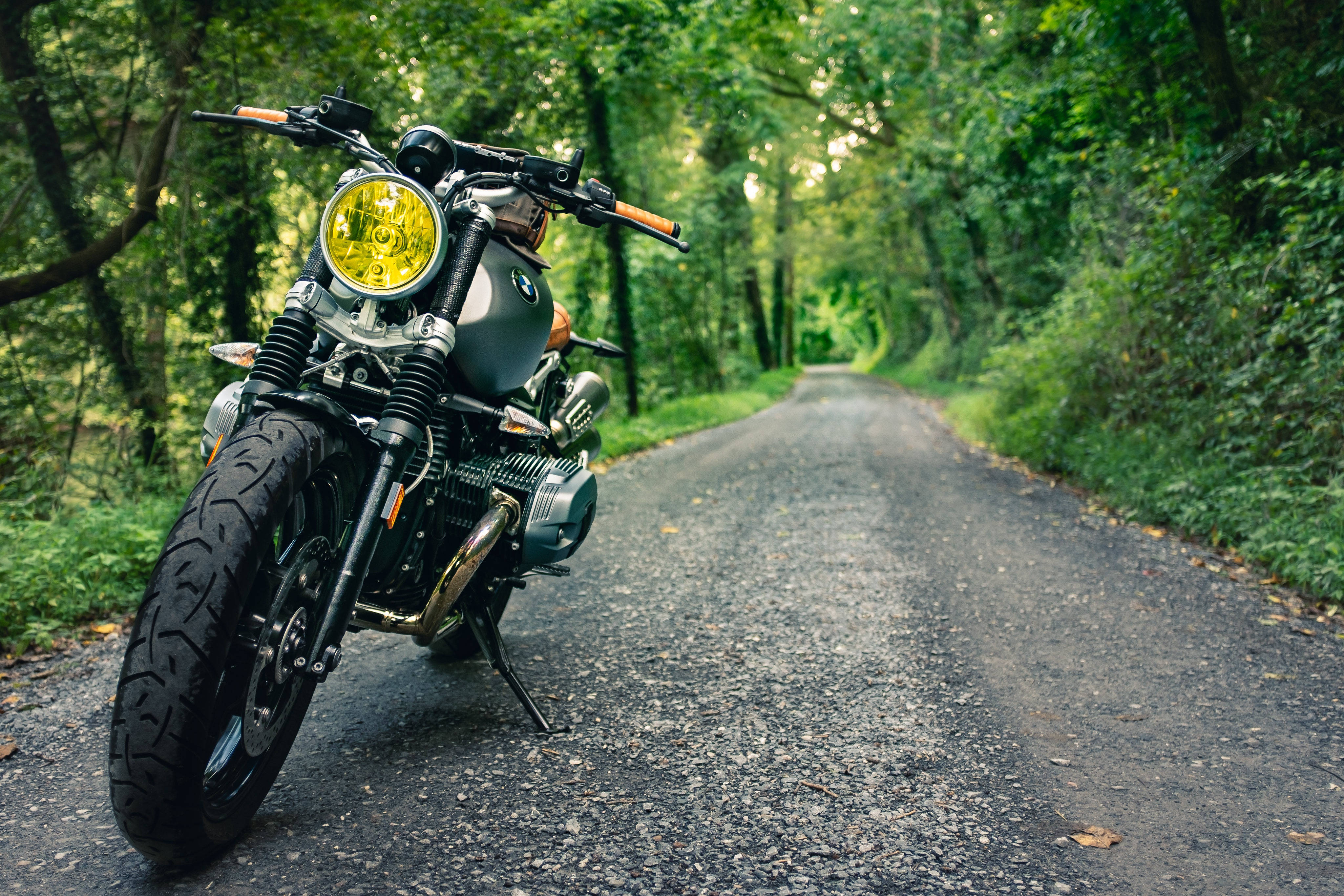 A vintage-style motorcycle with a bright yellow headlight parked on a gravel road winding through a lush green forest., 4K Desktop Wallpaper