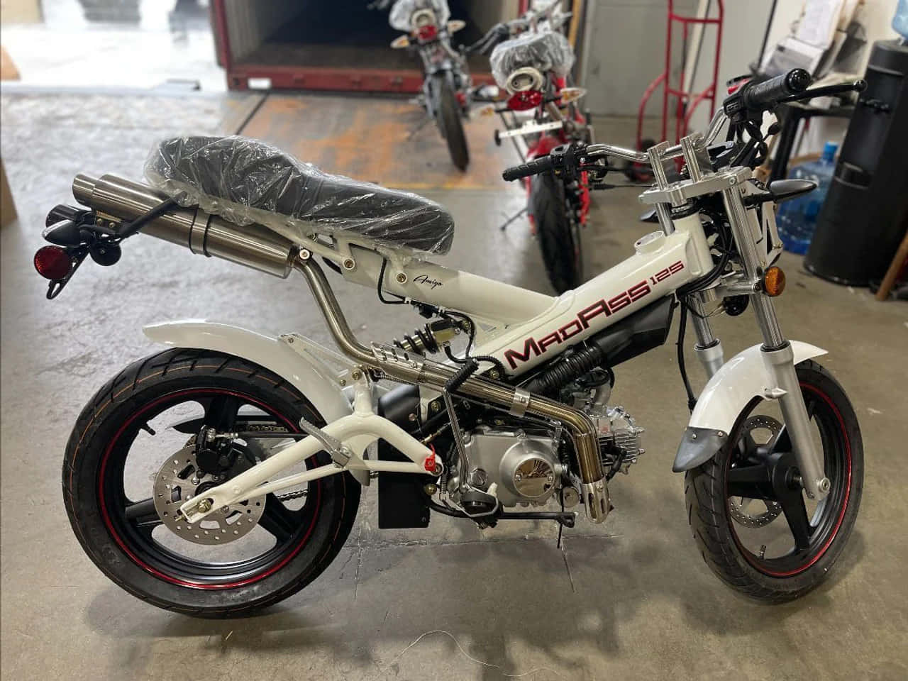 A white MadPass 125cc motorcycle with red accents and chrome exhaust sits in a garage with other bikes in the background., HD Desktop Wallpaper