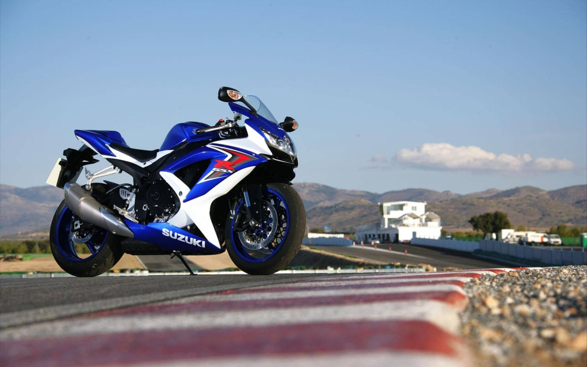 A blue and white Suzuki sport motorcycle parked on a racetrack with red and white curbing mountains and a clear sky in the background., Full HD Desktop Wallpaper