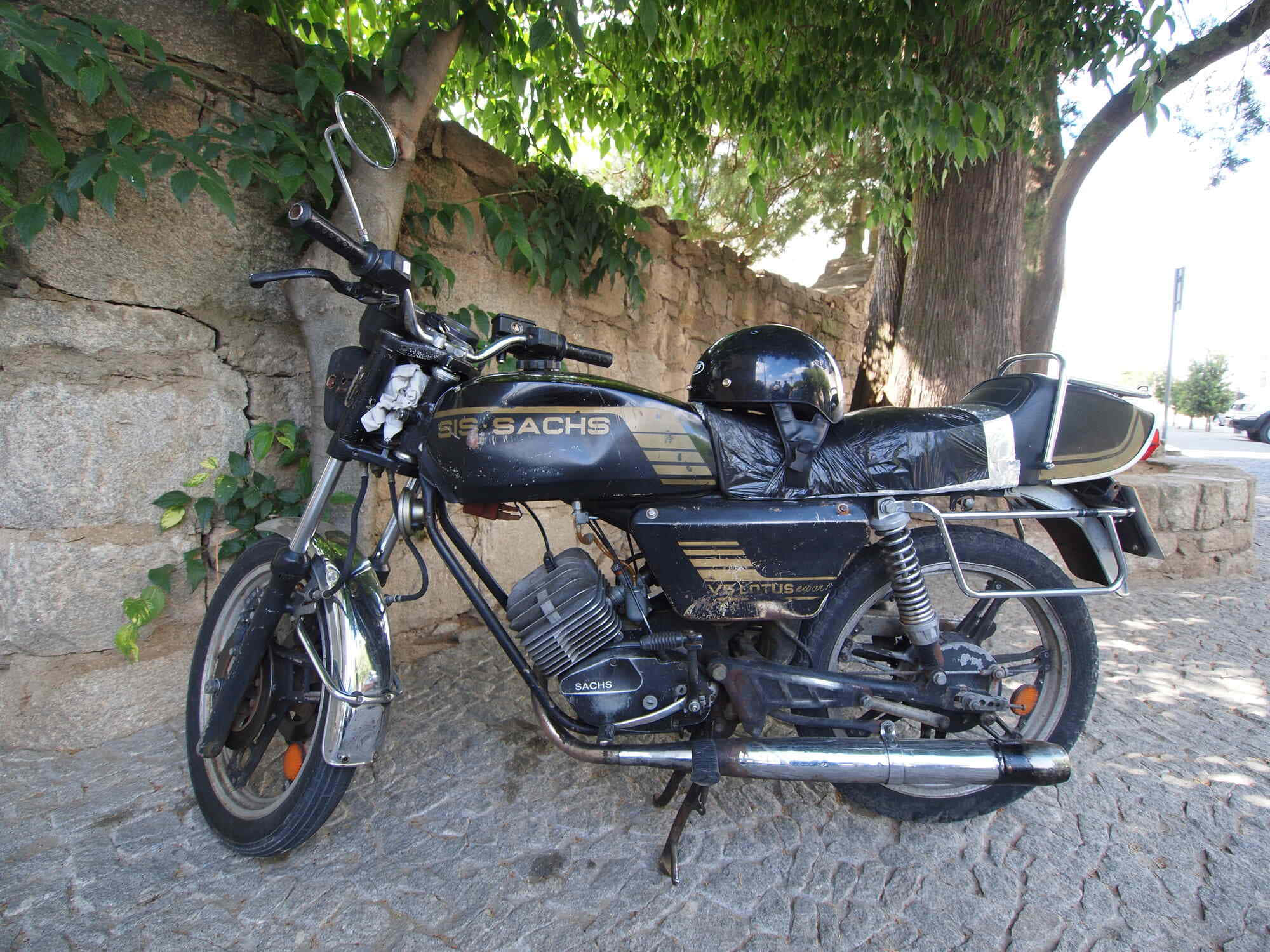 A vintage black Sachs motorcycle with chrome accents rests on a cobblestone street beside a stone wall and leafy tree., Full HD Desktop Wallpaper