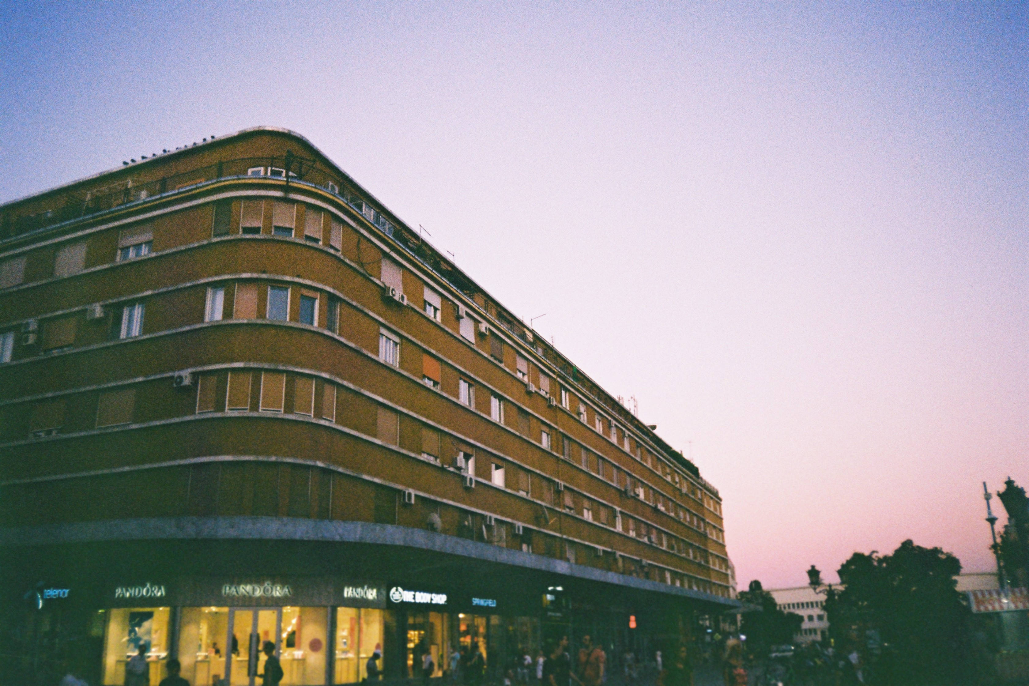 A low-angle view of a curved brick apartment building at dusk with warm storefront lights glowing against a soft pink and blue sky., 2K Desktop Wallpaper