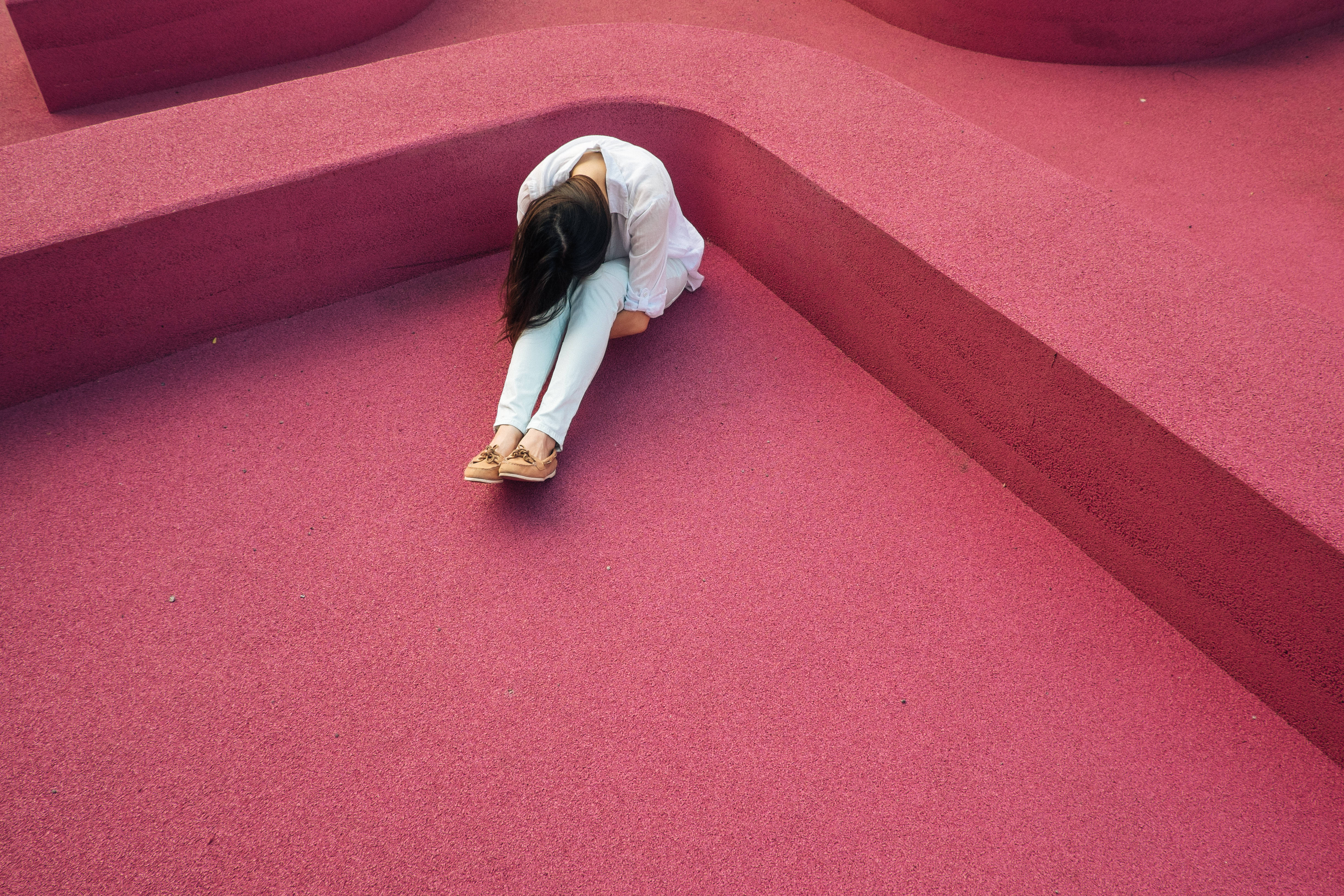 A person in white clothes sits curled on a vibrant pink textured surface, head bowed in quiet solitude., 4K Desktop Wallpaper