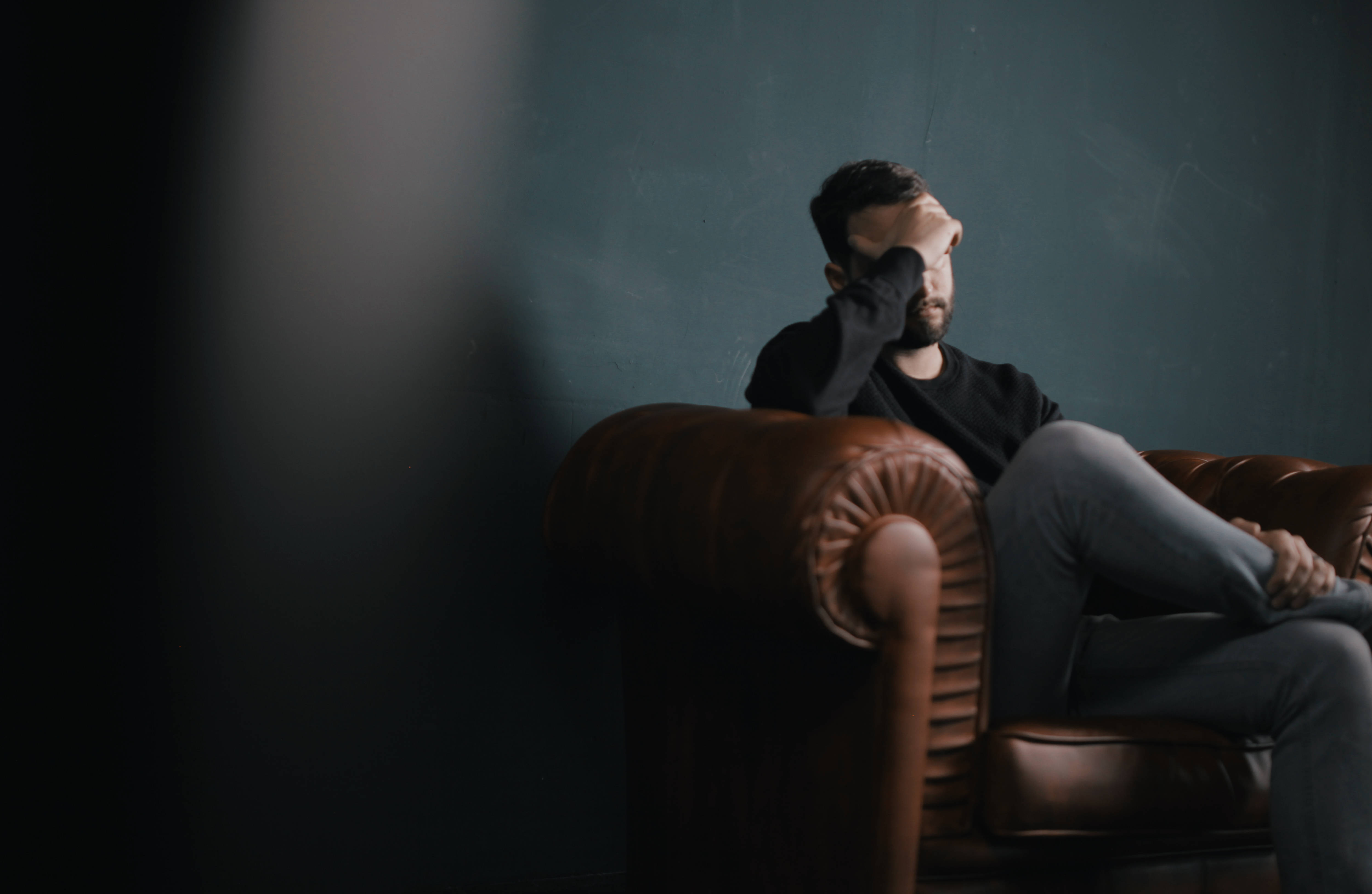 A man sits slumped in a leather armchair, hand over his face, in a dimly lit room with a dark teal wall., 4K Desktop Wallpaper