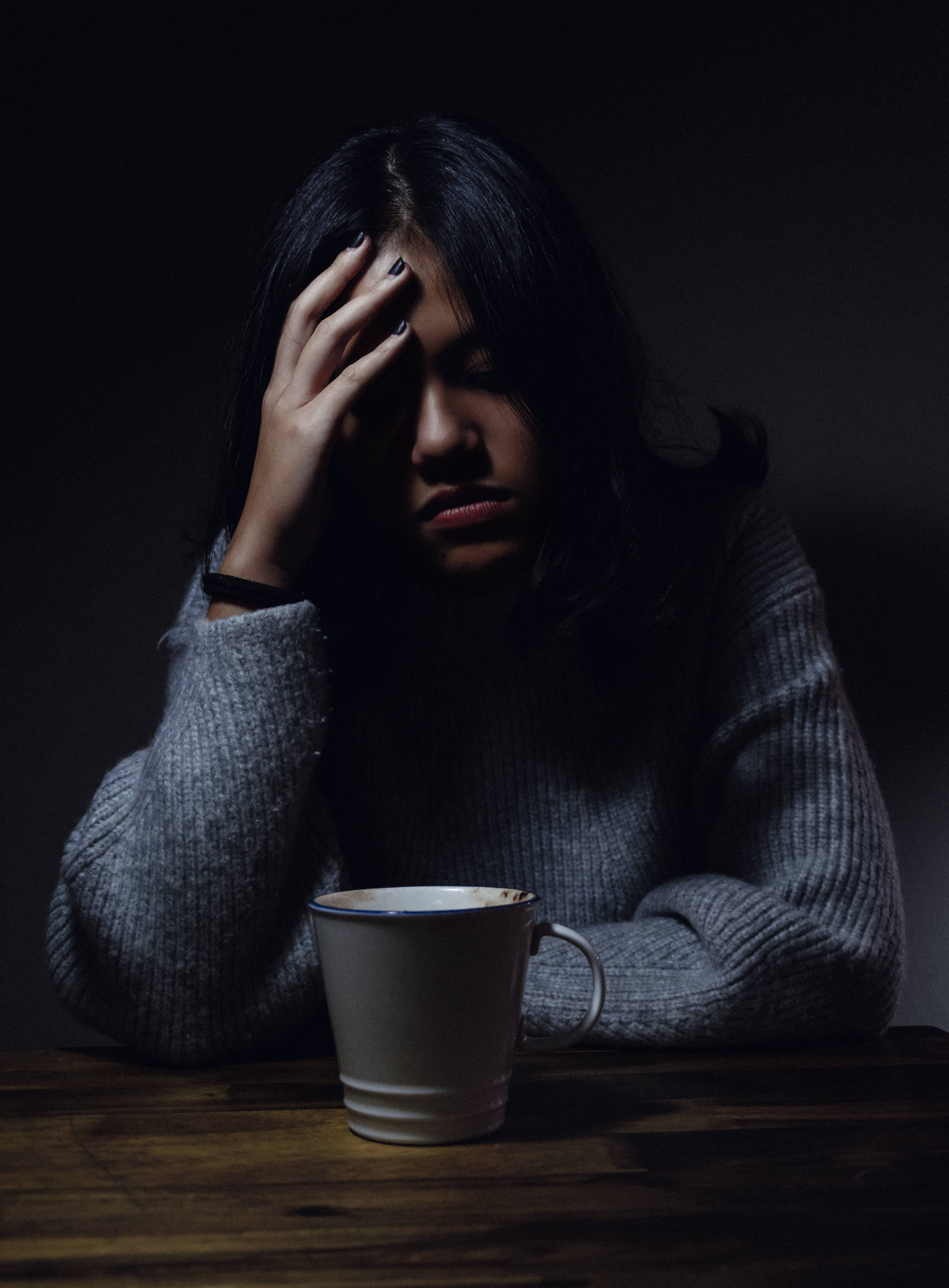 A woman in a gray sweater sits at a wooden table holding her head in her hand beside a white mug in dim lighting., 4K Phone Wallpaper