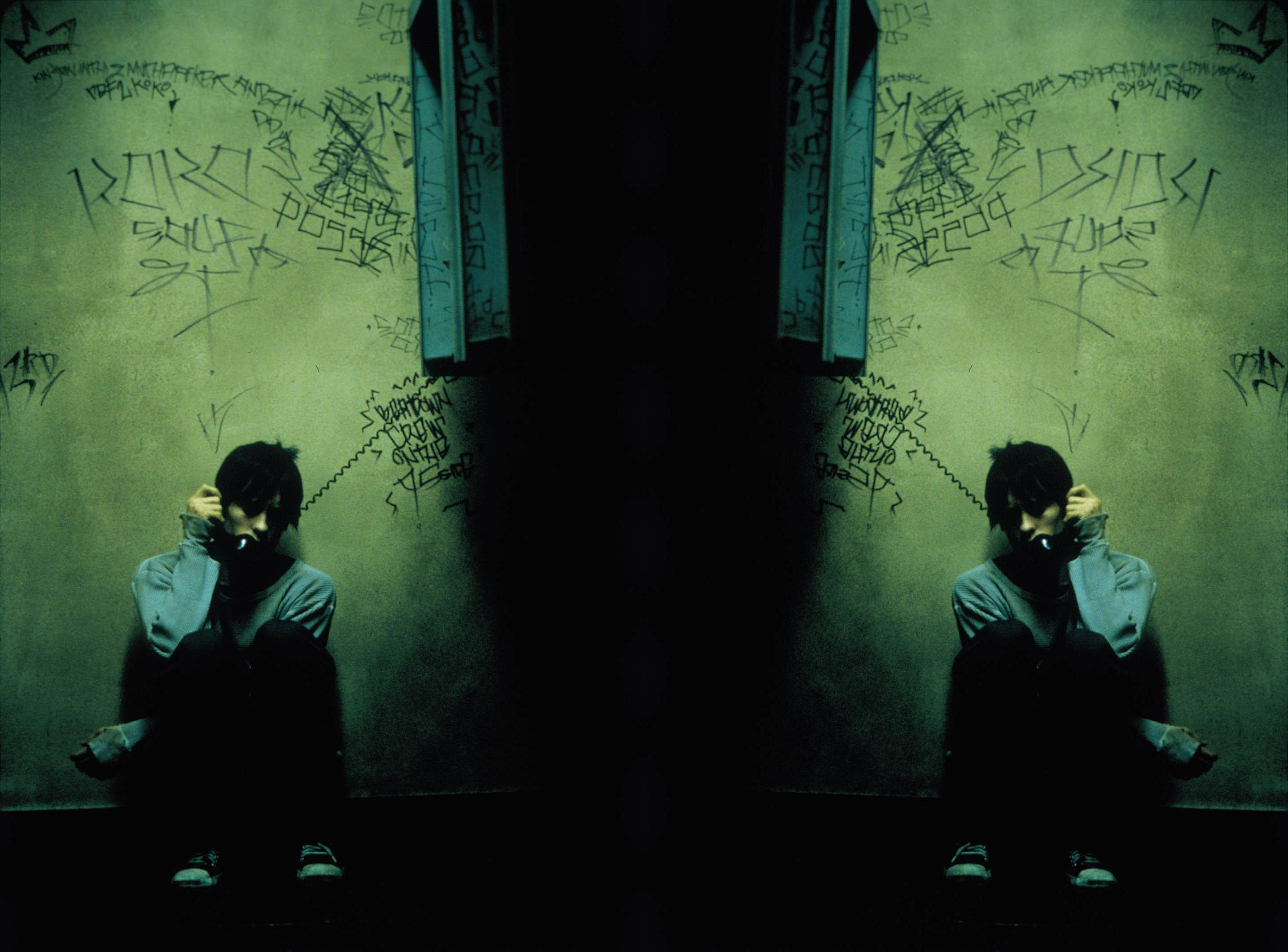 Two mirrored figures sit hunched in a dim green-lit room covered in graffiti, hands over ears in despair., 2K Desktop Wallpaper