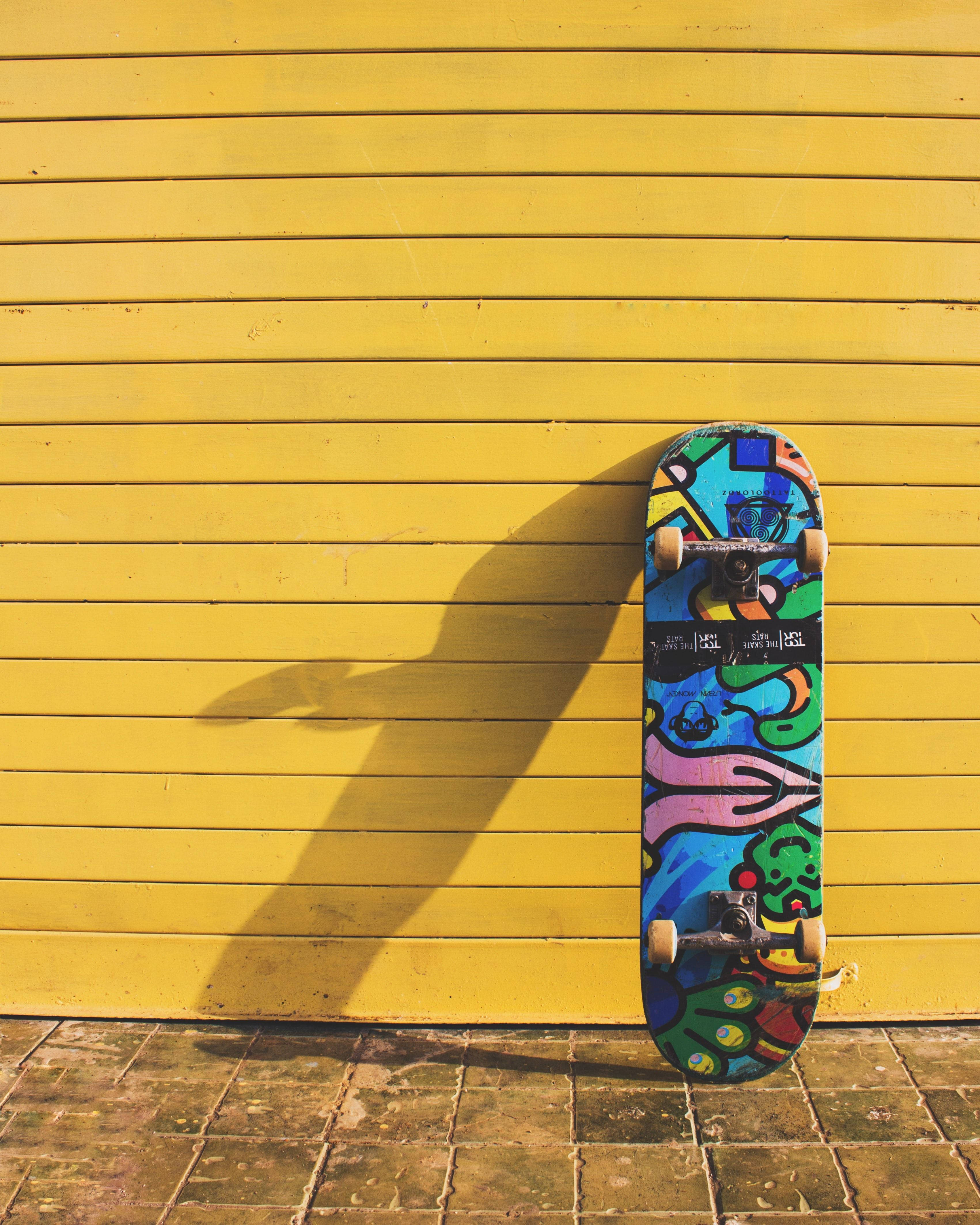colorful skateboard, bright yellow wooden wall, sunlit pavement, 4K Phone Wallpaper