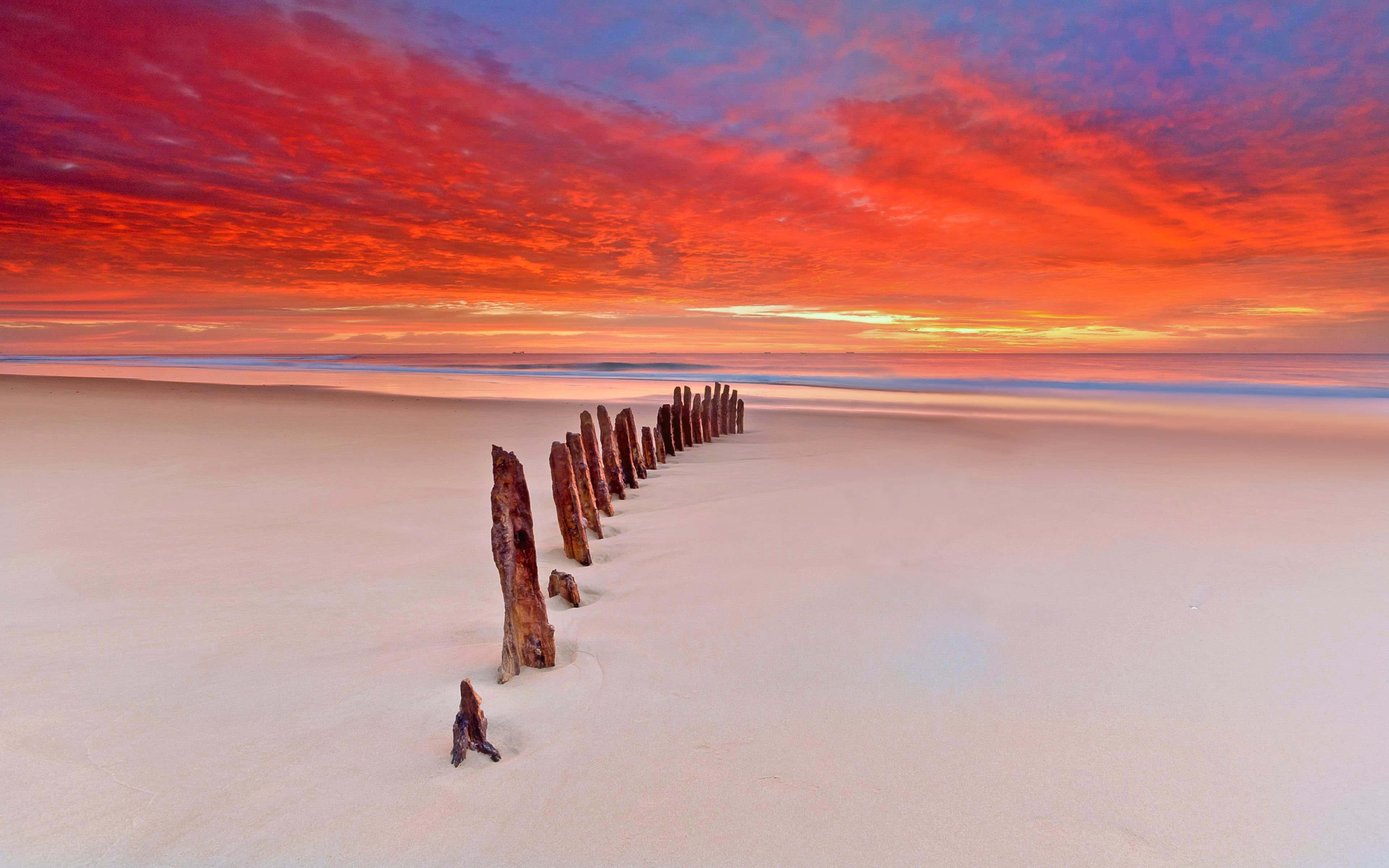 weathered wooden posts, pristine white sand beach, fiery red orange sunset sky, 4K Desktop Wallpaper