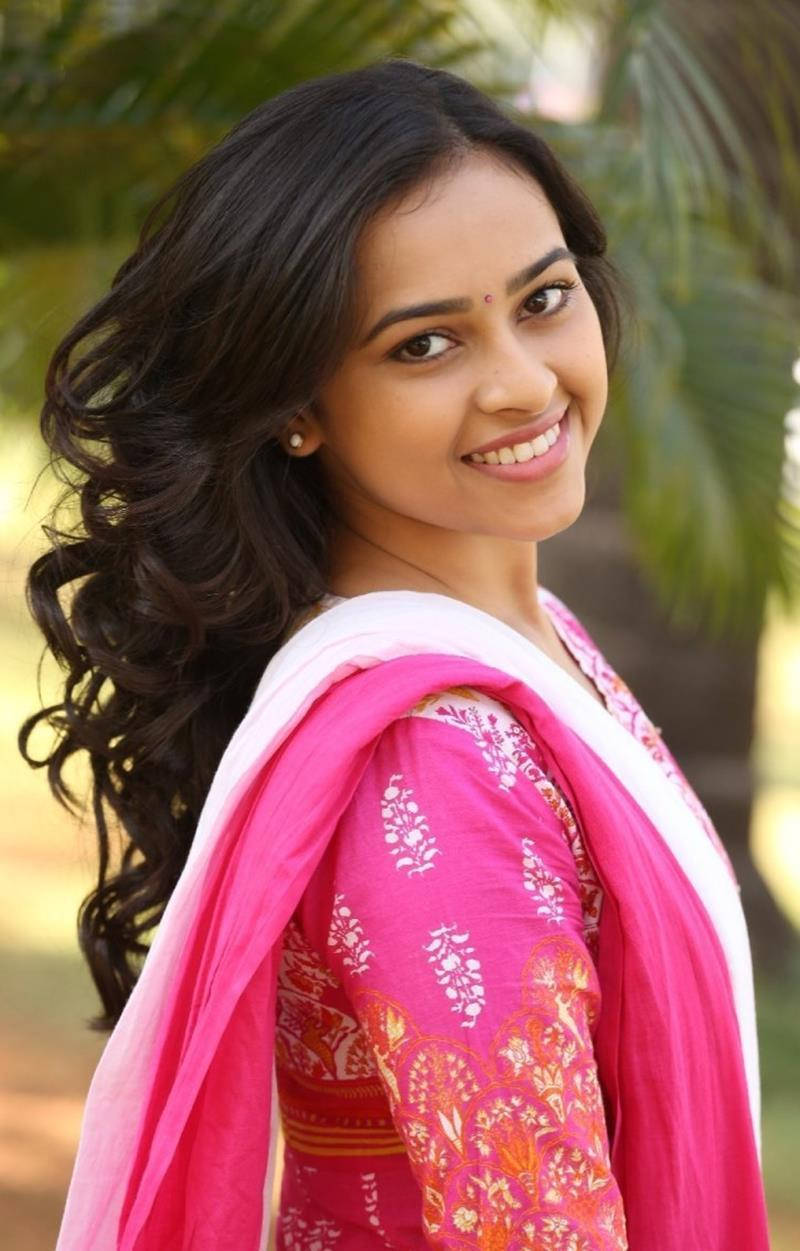 Sri Divya, vibrant pink and white patterned traditional outfit, blurred green palm tree background, SD Phone Wallpaper