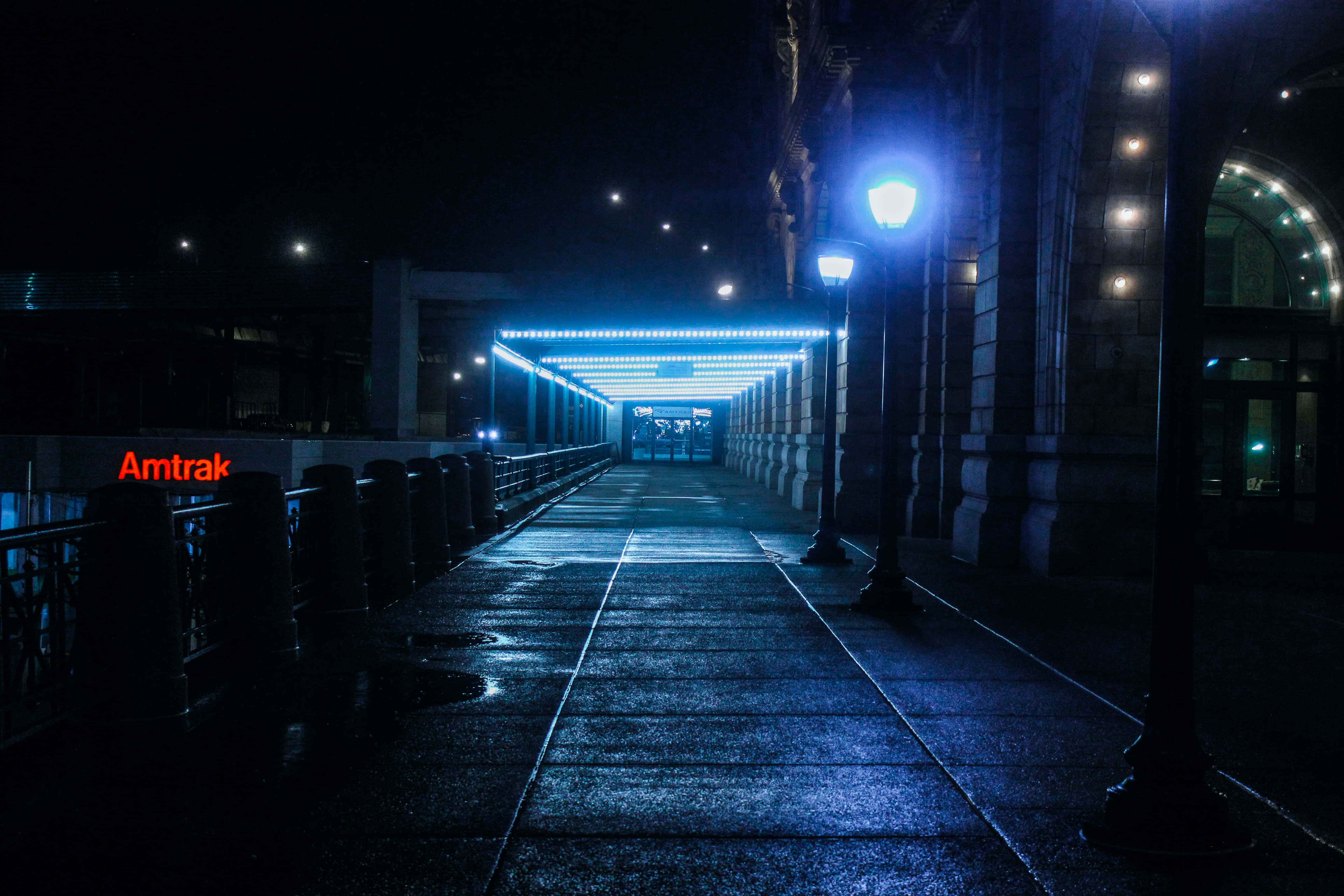 Amtrak station entrance, glowing blue lights, wet nighttime walkway, 5K Desktop Wallpaper