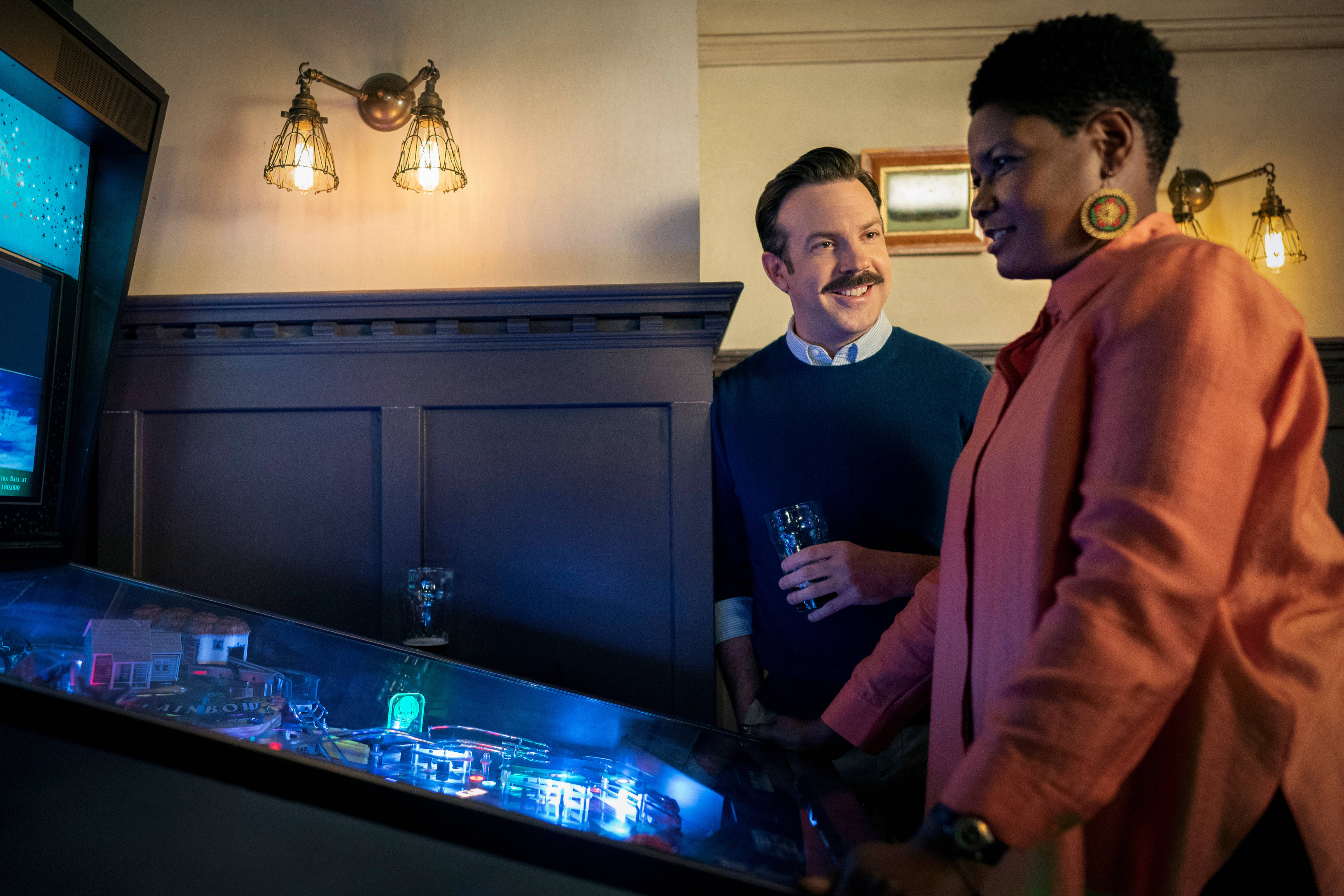 Ted Lasso, glowing pinball machine, warmly lit vintage arcade, 4K Desktop Wallpaper