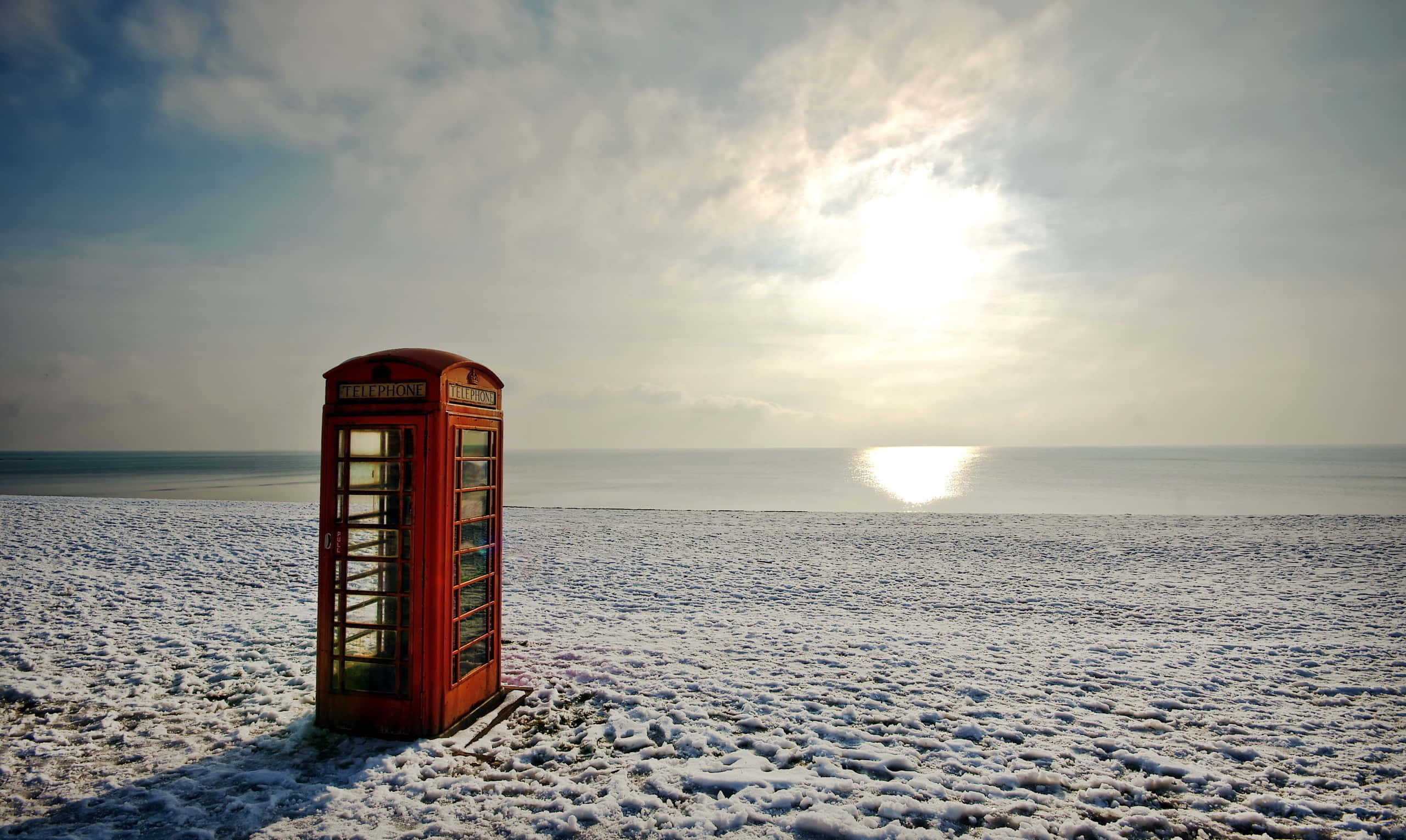 red British telephone box, snow-covered beach, sun glows over the calm sea, 2K Desktop Wallpaper