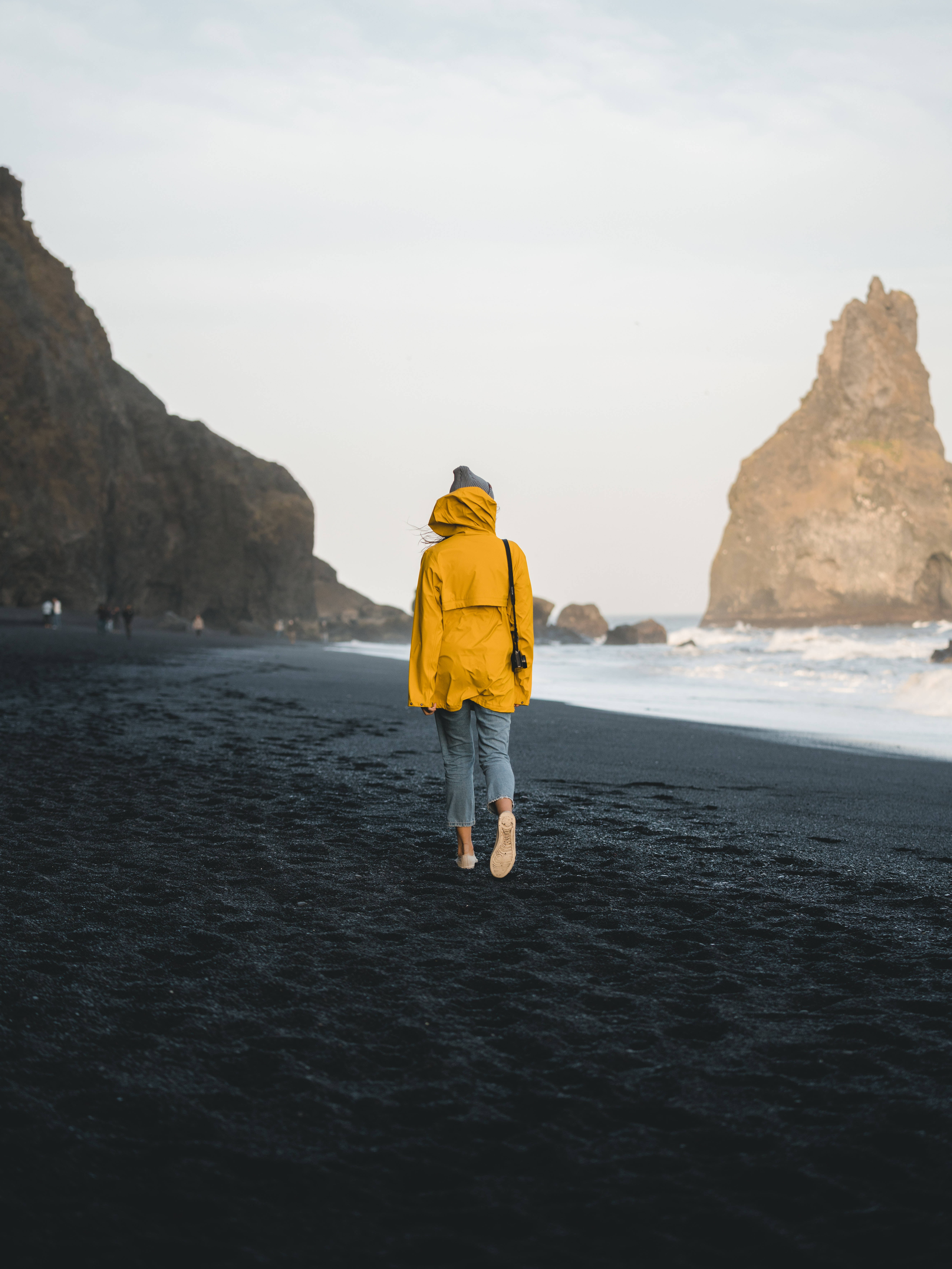 bright yellow raincoat, stark black volcanic beach, towering cliffs, 5K Phone Wallpaper